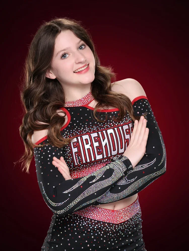 Young girl in a black cheerleading costume with red and white accents, rhinestones, and the word 'FIREHOUSE' across the chest, posing with arms crossed against a red gradient background.