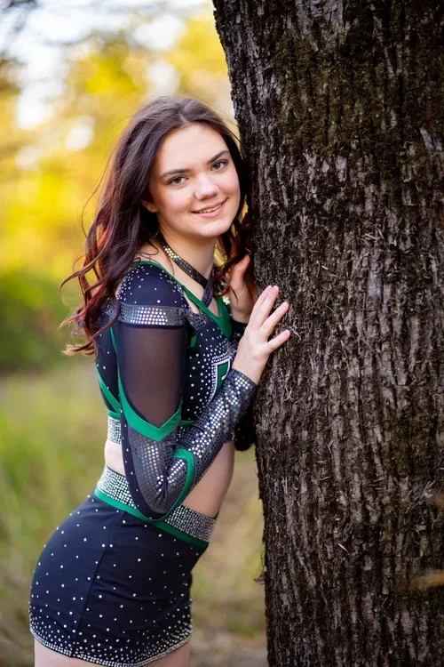 Young woman outdoors in a cheerleading uniform, standing next to a large tree, smiling at the camera.