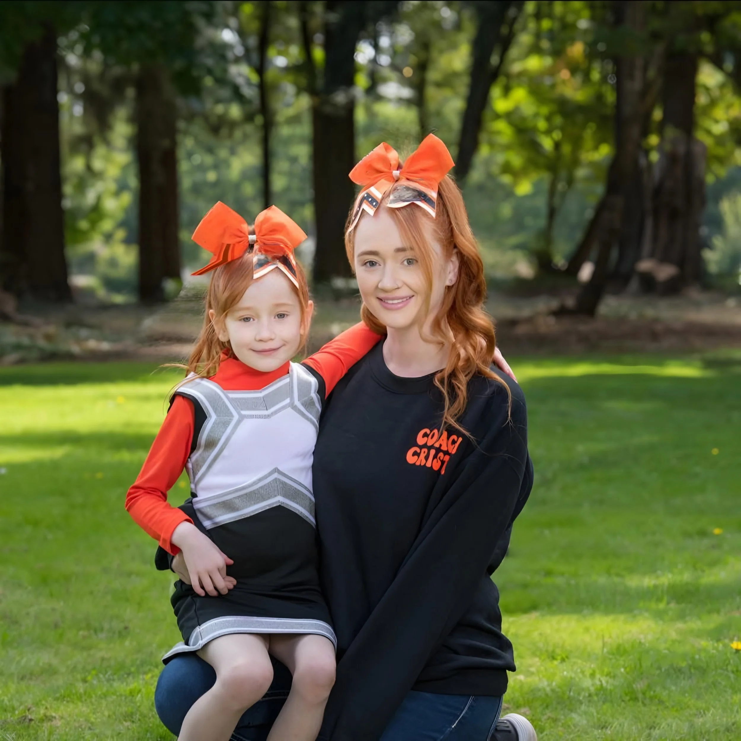A woman and a young girl sit on a grassy field in a park, both wearing cheerleading outfits with matching orange bows in their hair. The woman is smiling and wearing a black shirt with red lettering that says 'COACH CRIST.' The girl is sitting on the woman's lap, smiling, and wearing a cheerleading uniform with red, black, and gray colors.