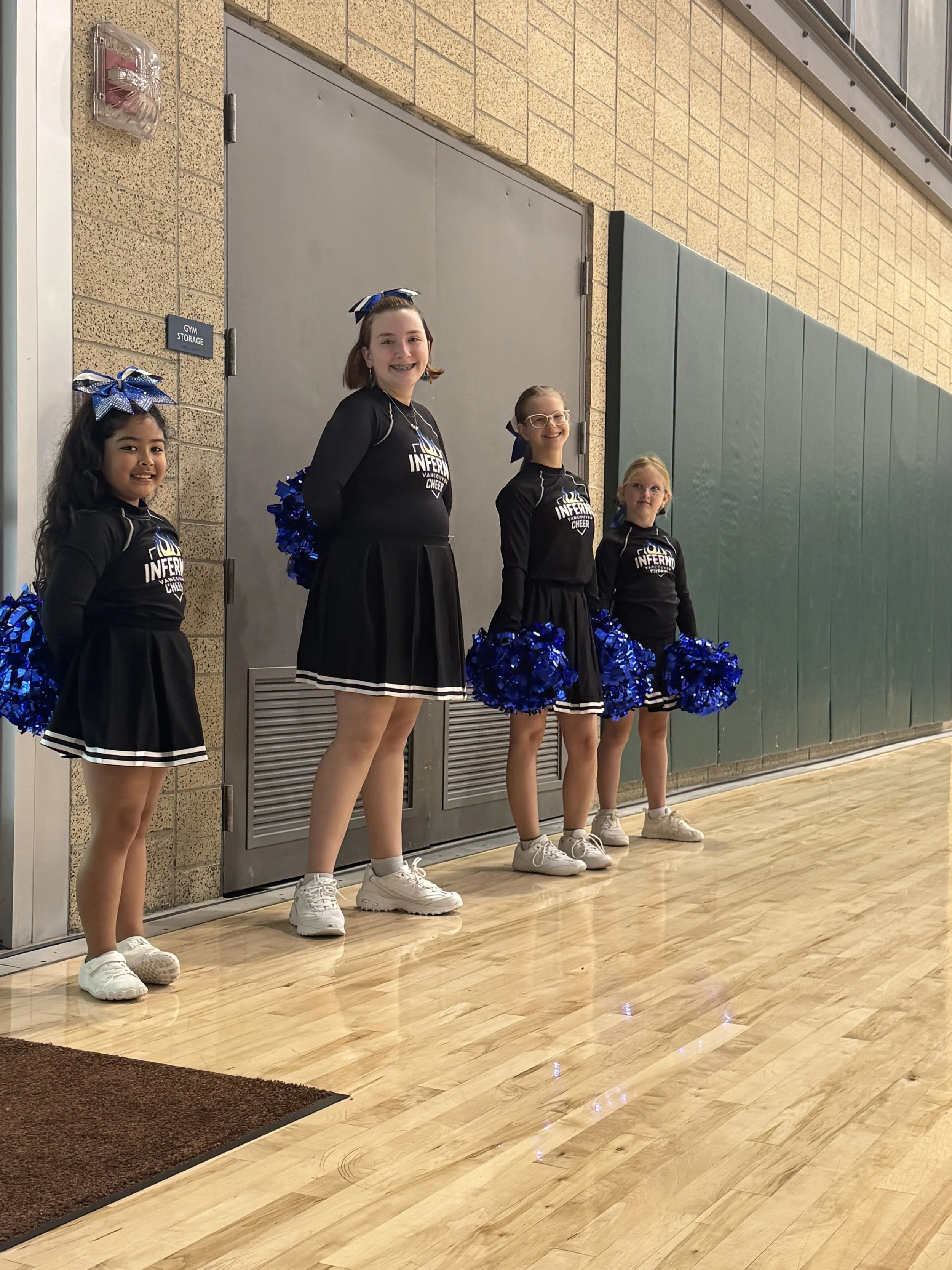 Four young cheerleaders standing side by side inside a gymnasium, holding blue pom-poms, wearing matching black uniforms with the words "Inferno Cheer" and a flame logo, smiling.