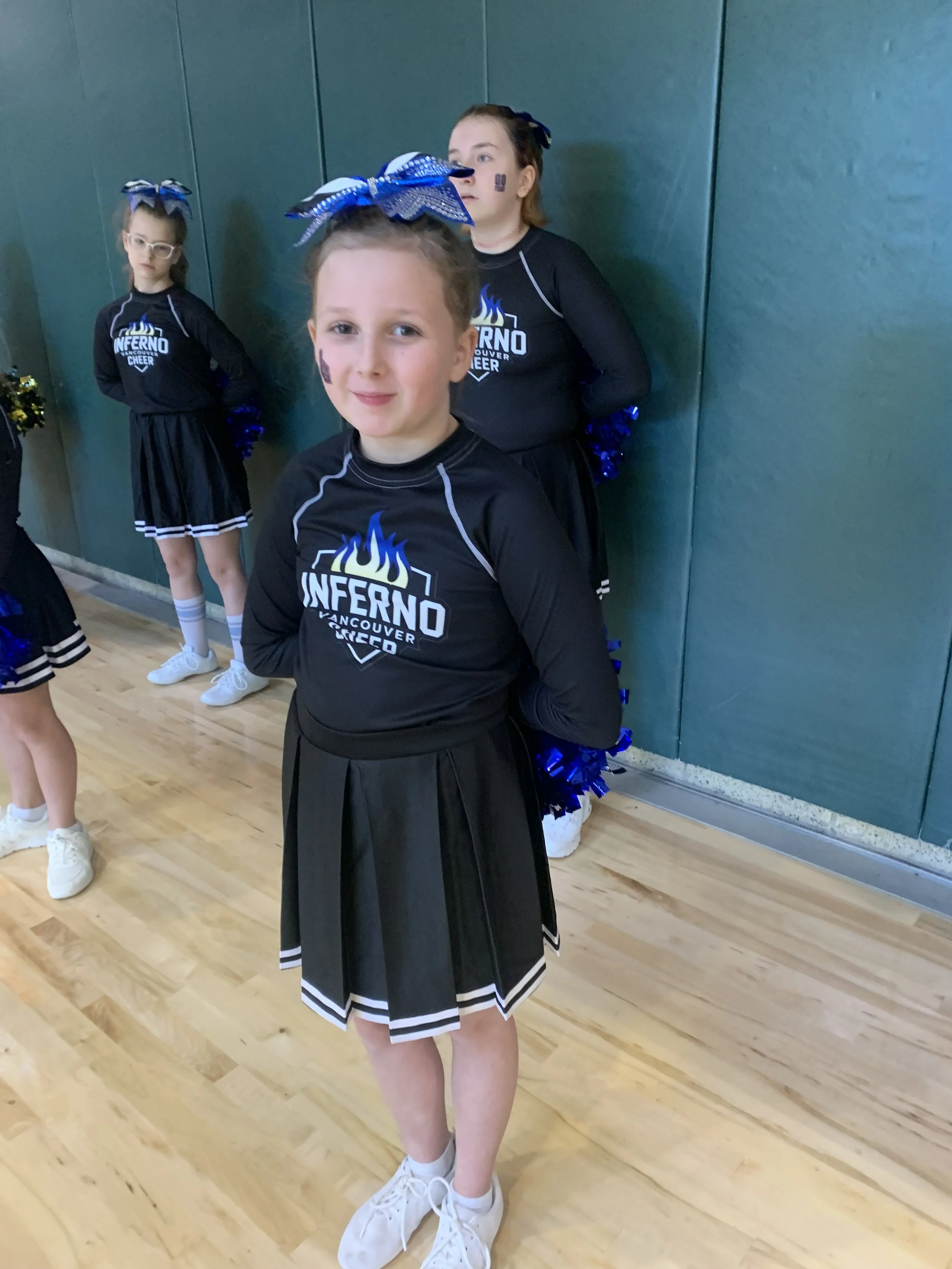 Three cheerleaders in black uniforms with blue and white pom-poms standing against a green wall in a gym. The girl in front has a blue bow, face paint, and white sneakers. The two girls behind have blue bows, face paint, and glasses.