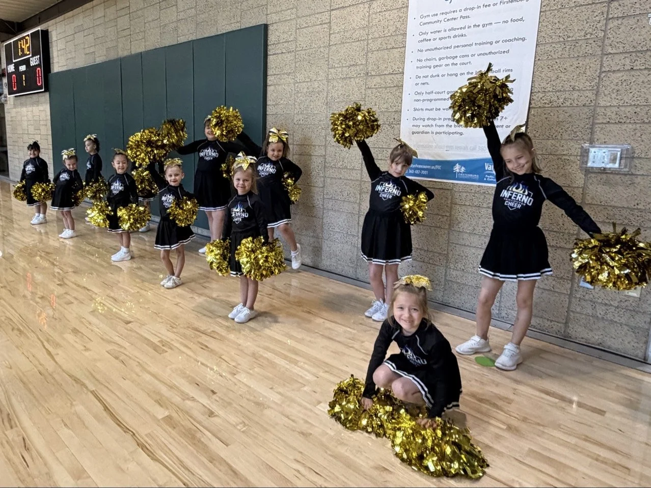 Group of young cheerleaders in black uniforms with gold pom-poms, standing in a line in an indoor gymnasium, posing for a photo.
