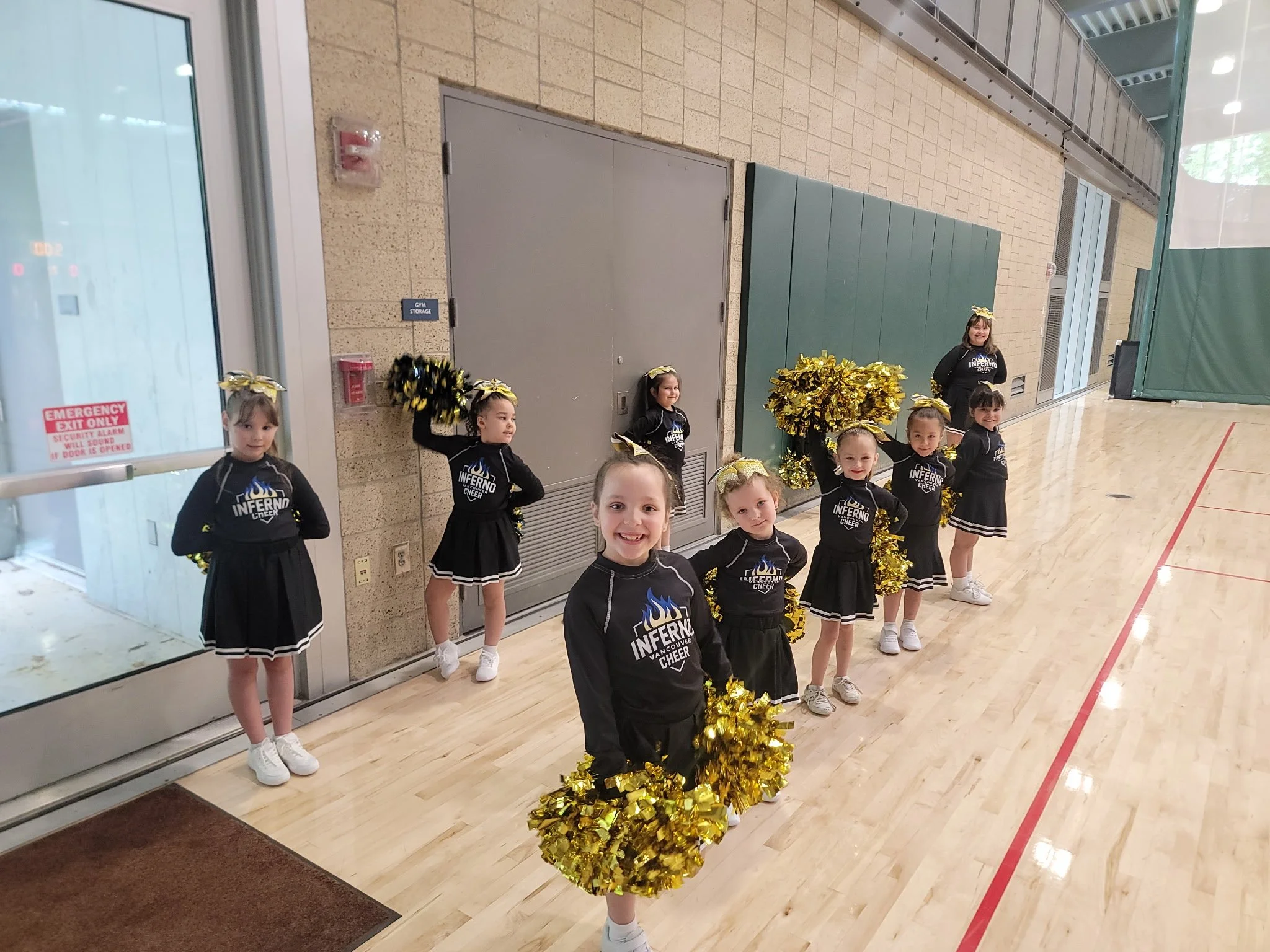 A group of young cheerleaders wearing black uniforms with gold pom-poms, standing inside a gymnasium near a wall with a fire alarm, a door, and a sign that says 'Gym Storage.'