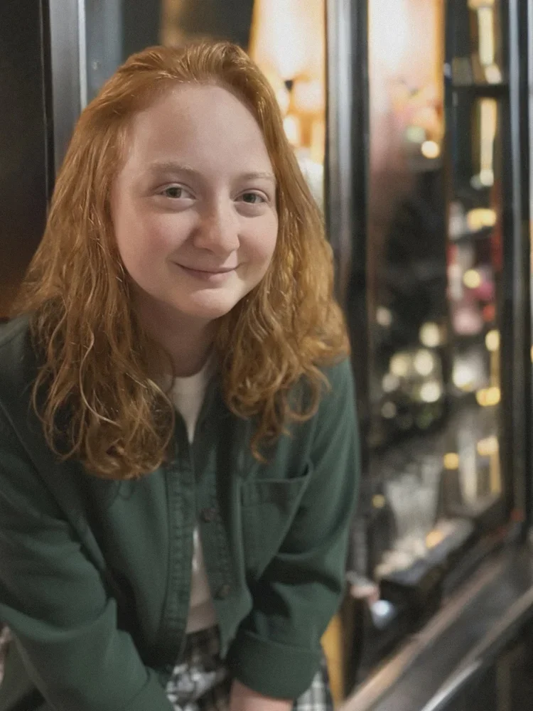 A young woman with curly red hair smiling in front of a vending machine in a cozy, warmly lit setting.