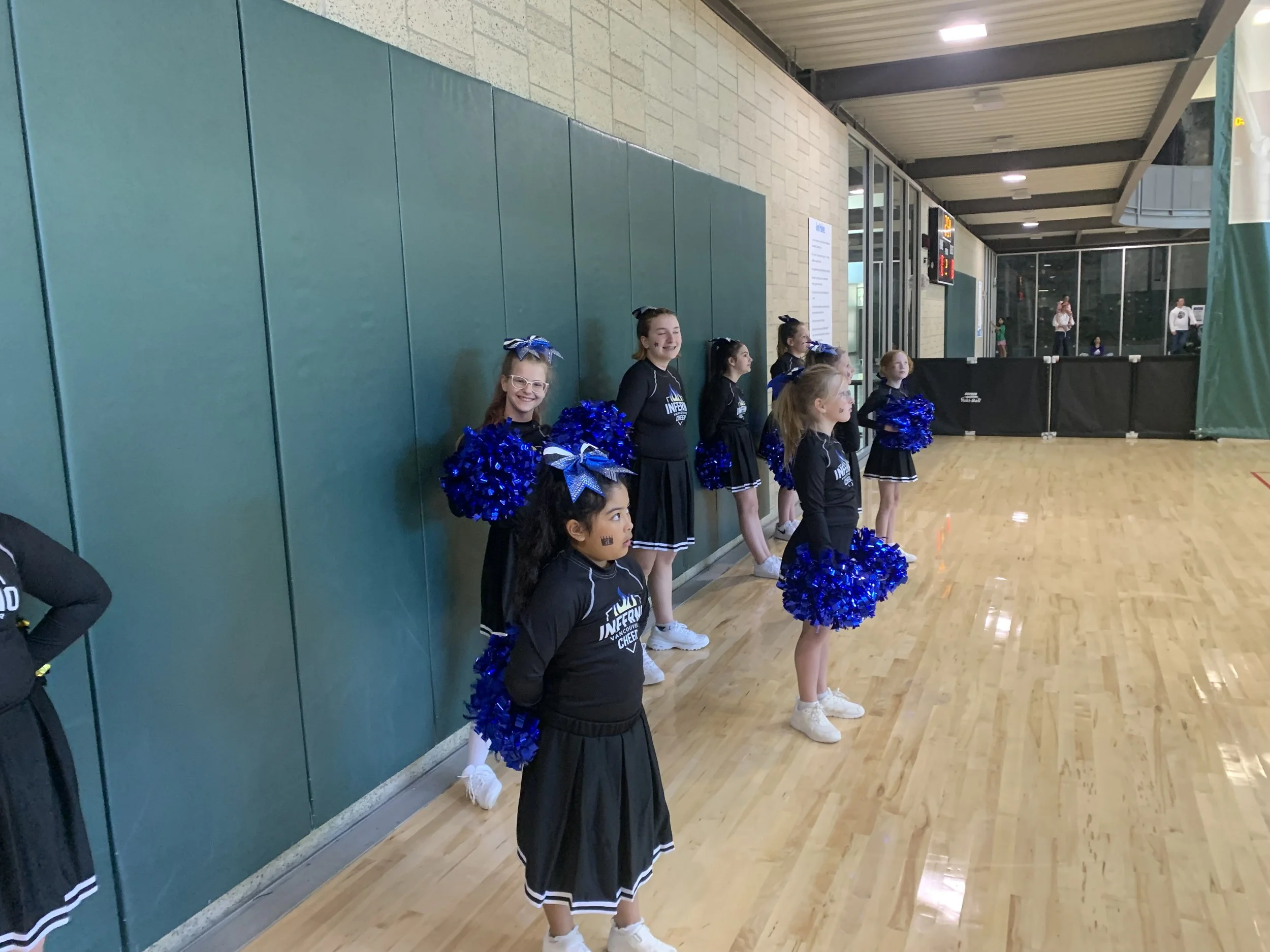 A group of cheerleaders standing in a line inside a gymnasium, with some holding blue pom-poms and dressed in black cheerleading uniforms with blue bows in their hair, smiling and preparing for a performance.