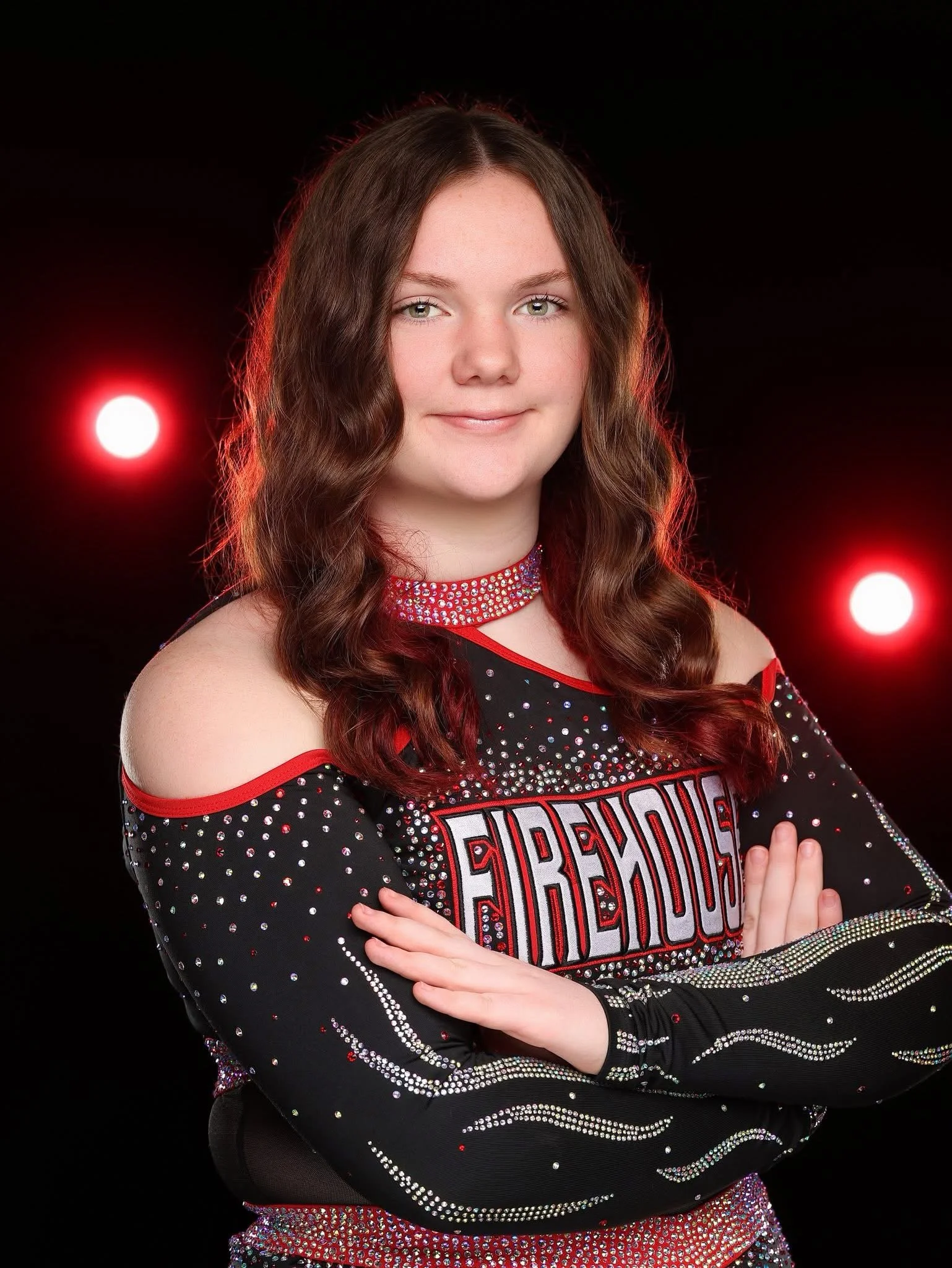 A young woman with brown wavy hair in a cheerleading uniform with rhinestones, standing with arms crossed against a black background with red lights.