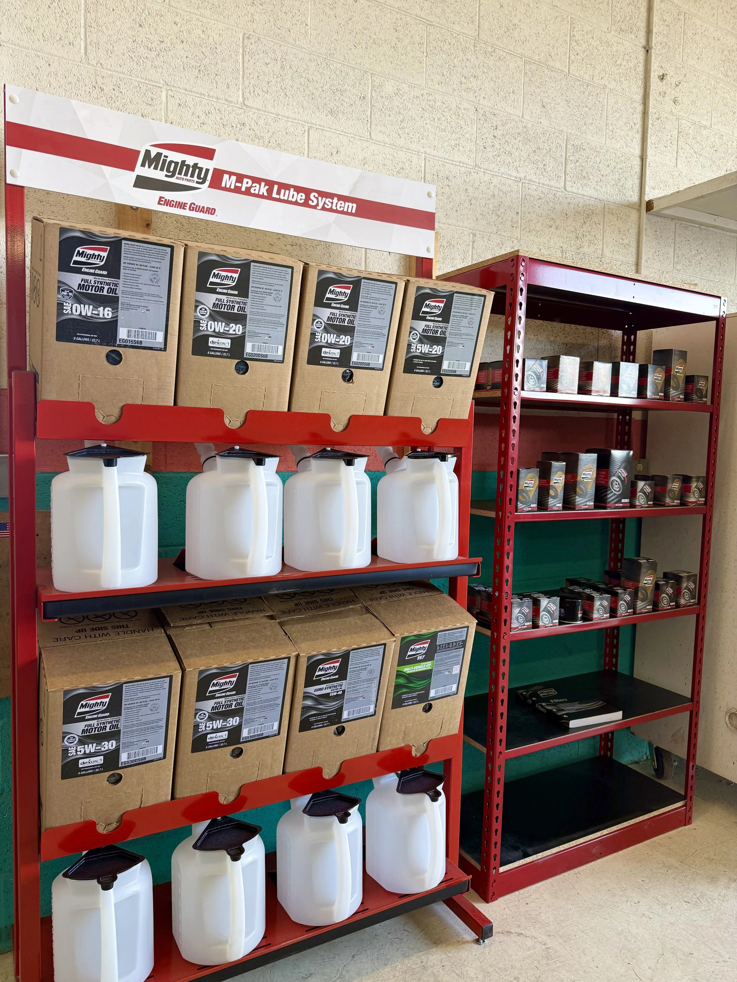 Red industrial shelf holding boxes of Mighty engine oil bottles and white plastic spray bottles, near a beige wall with a sign reading "Mighty M-Pak Lube System".