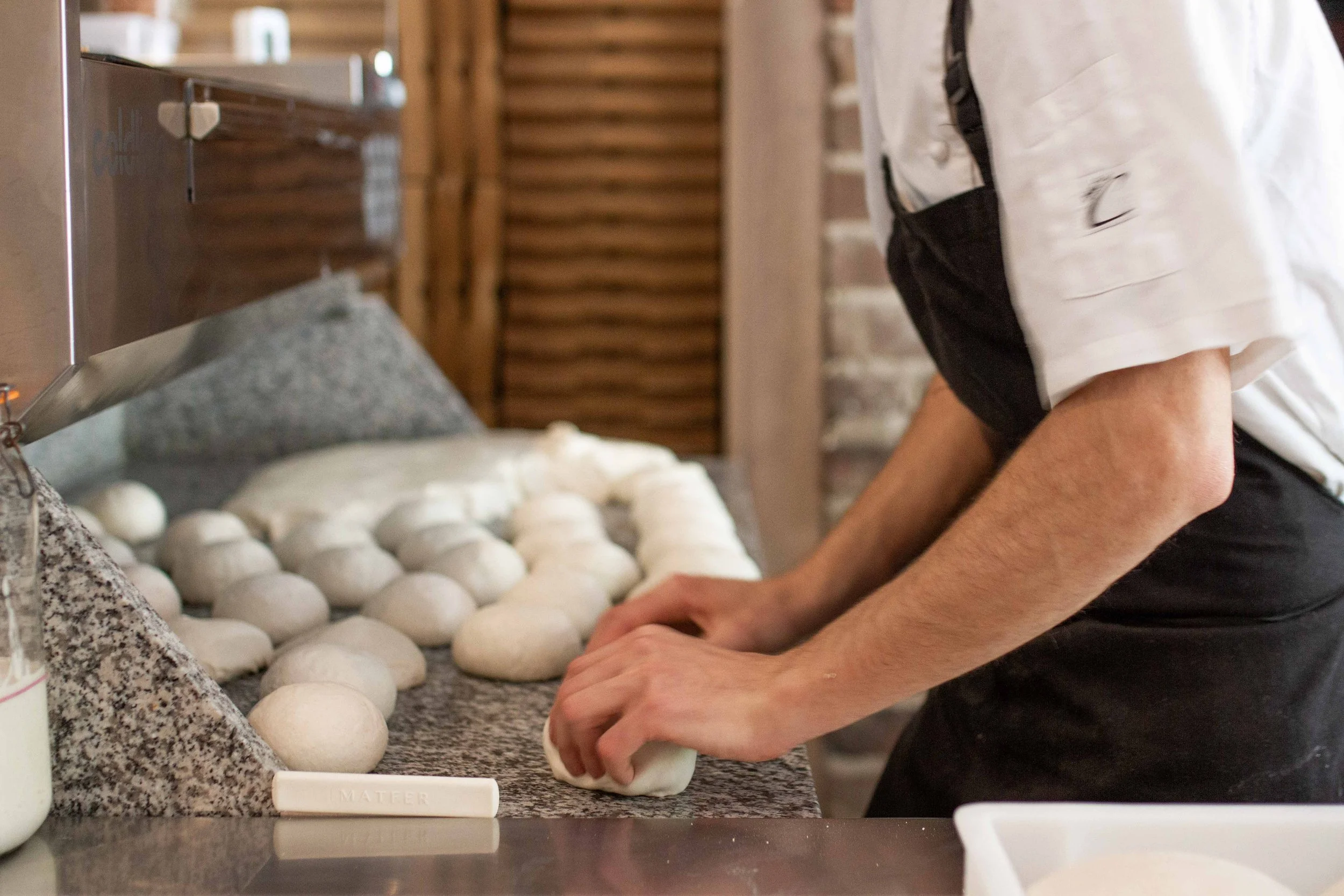 Person shaping dough into balls on a granite countertop in a bakery kitchen.