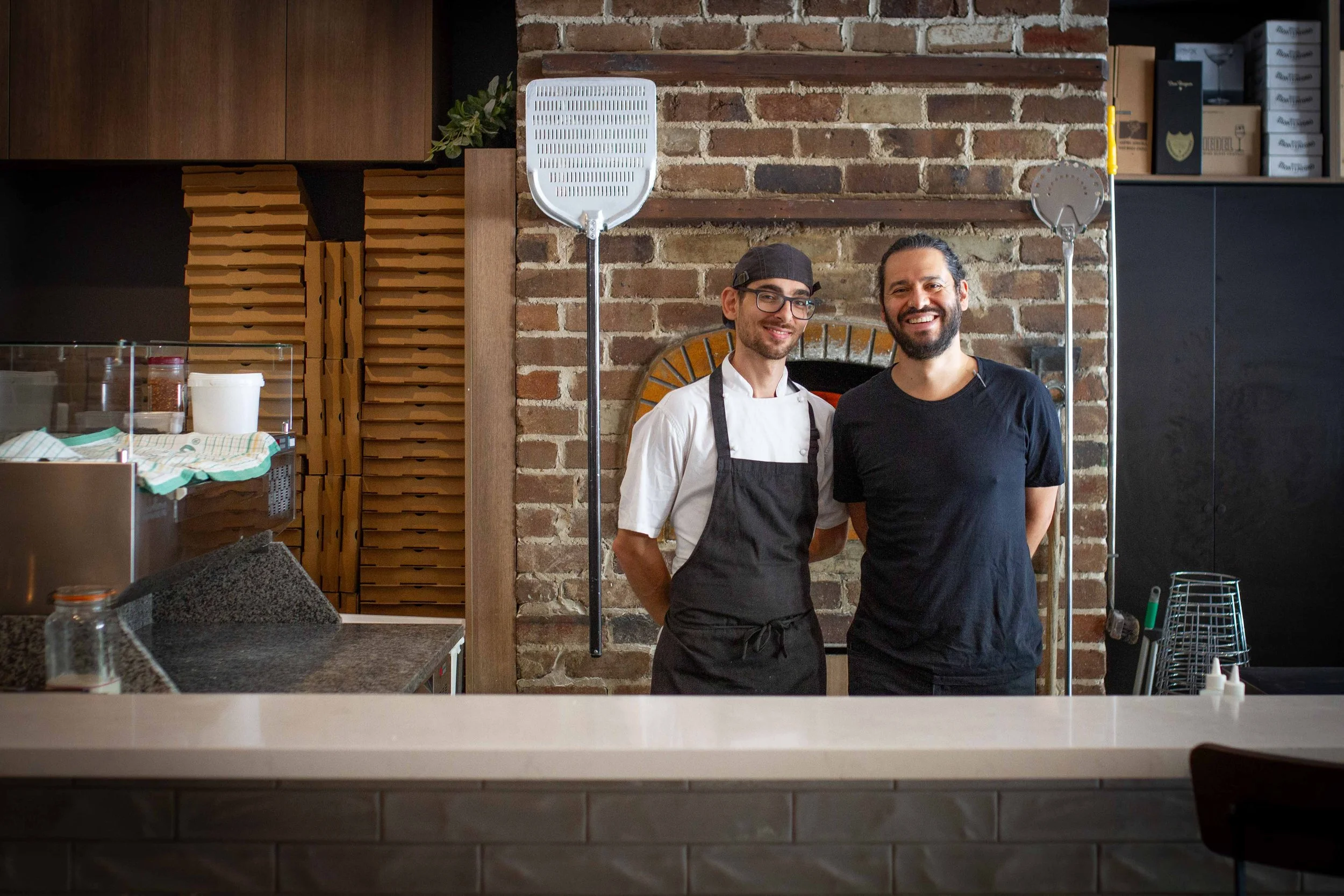 Stefano and Mauricio smiling inside a restaurant kitchen. One is wearing a black apron and white shirt, and the other is dressed in black. They stand in front of a brick wall and are flanked by pizza oven paddles.