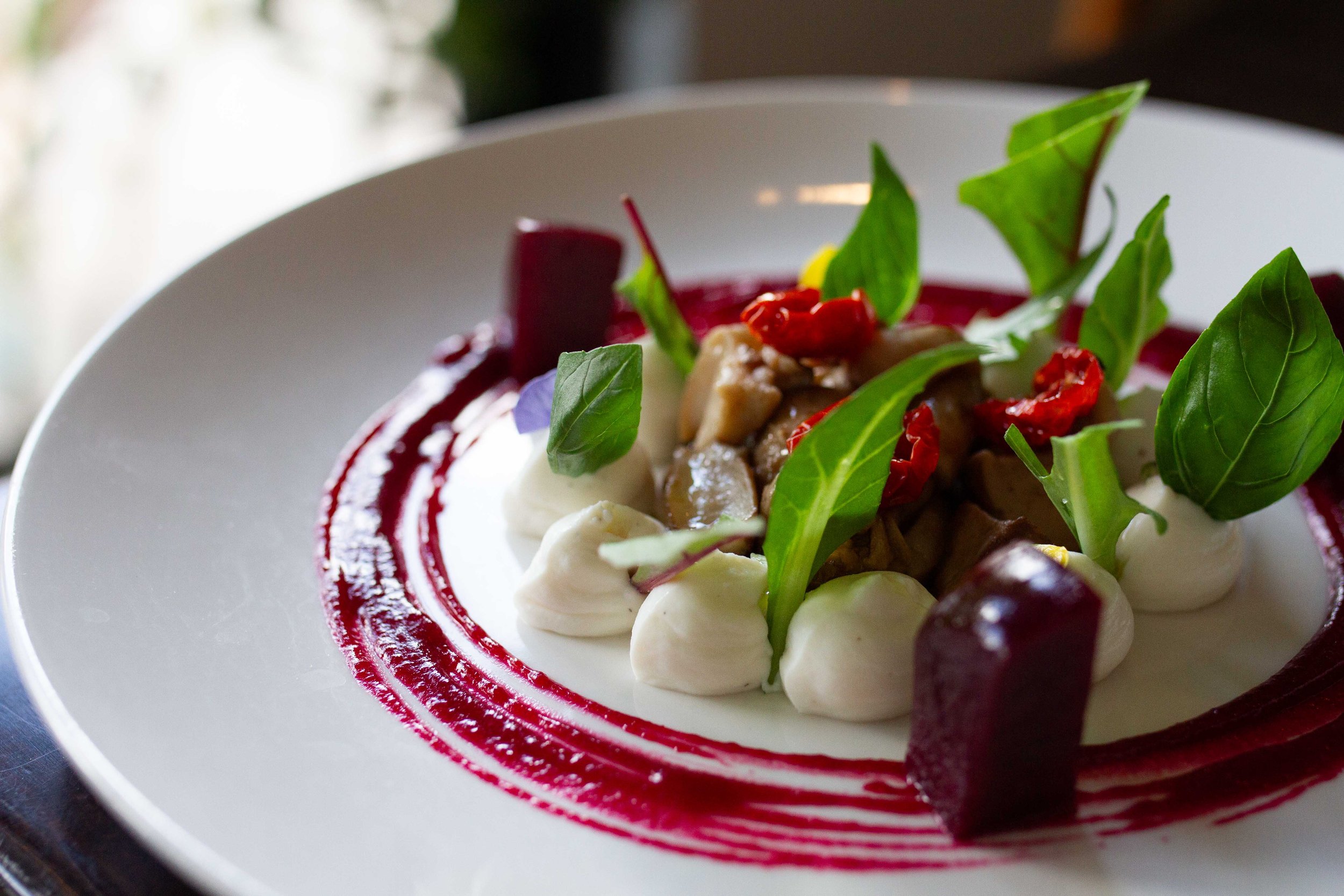 A plated gourmet dish with dollops of white sauce, fresh green herbs, red beets, and a brown protein on a white plate with a red sauce swirl.
