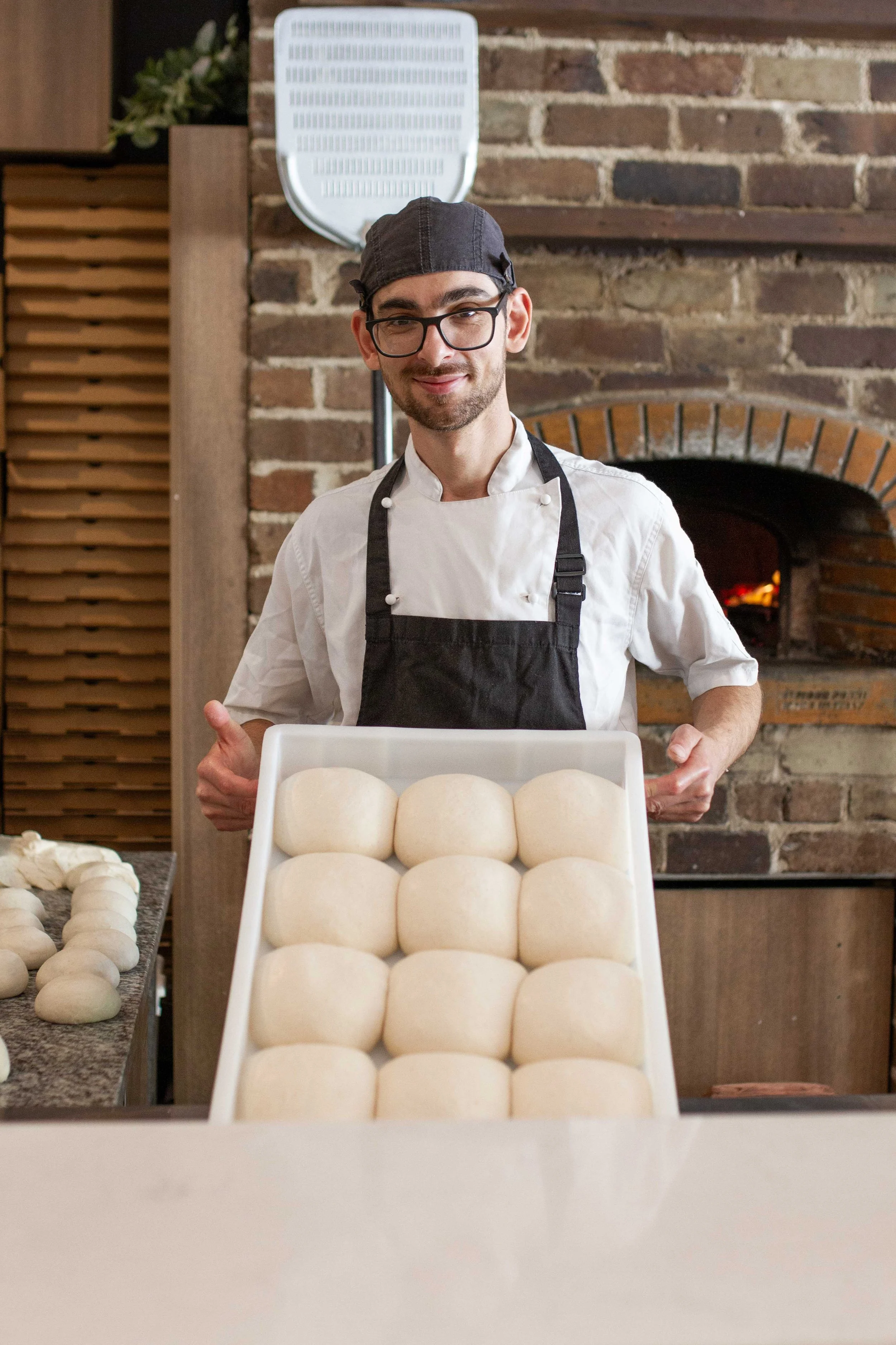 Stefano Spataro in a white chef's coat and black apron holding a tray of round, uncooked bread dough balls in a bakery kitchen with a brick oven in the background.
