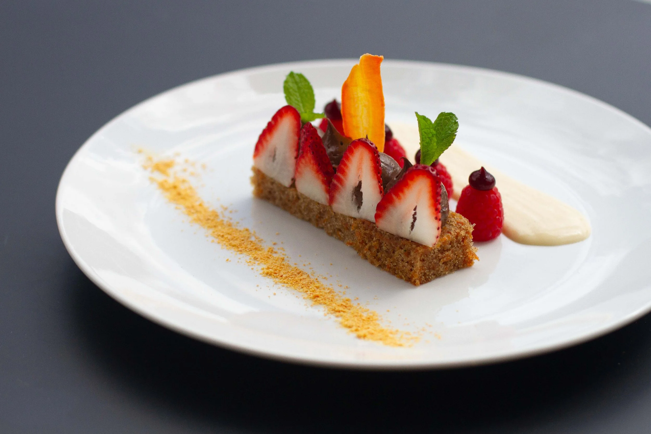 A slice of fruit tart on a white plate, decorated with strawberries, raspberries, a slice of orange, a mint leaf, and a dollop of cream, with a dusting of golden powder on the plate.