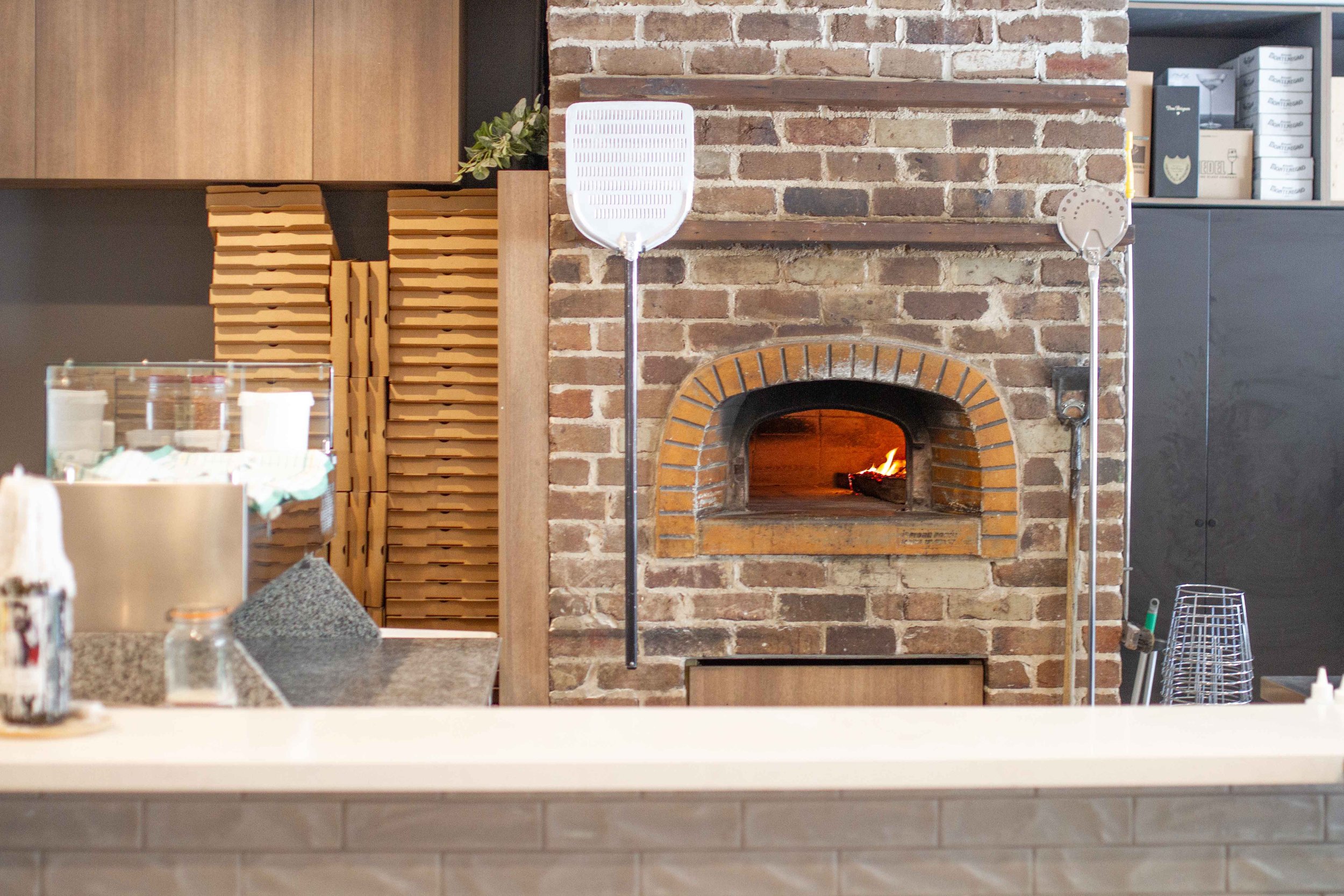 Indoor scene of a kitchen or bakery with a brick oven with a fire inside, surrounded by a brick wall. To the left of the oven, there are two modern heat lamps, and to the right, there are some kitchen tools and storage containers. The counter in the 
