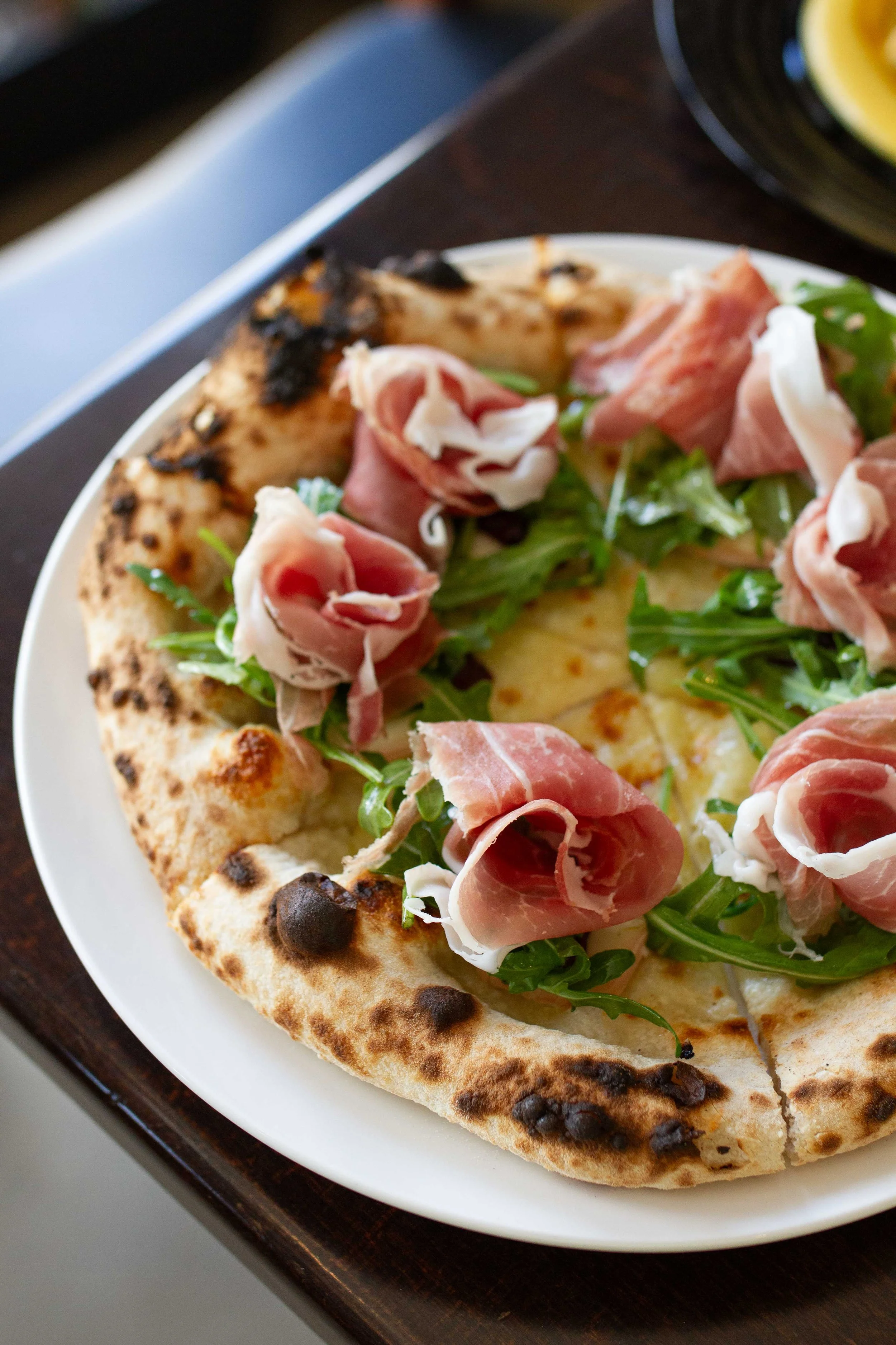 Close-up of a pizza with prosciutto, arugula, and melted cheese on a white plate.