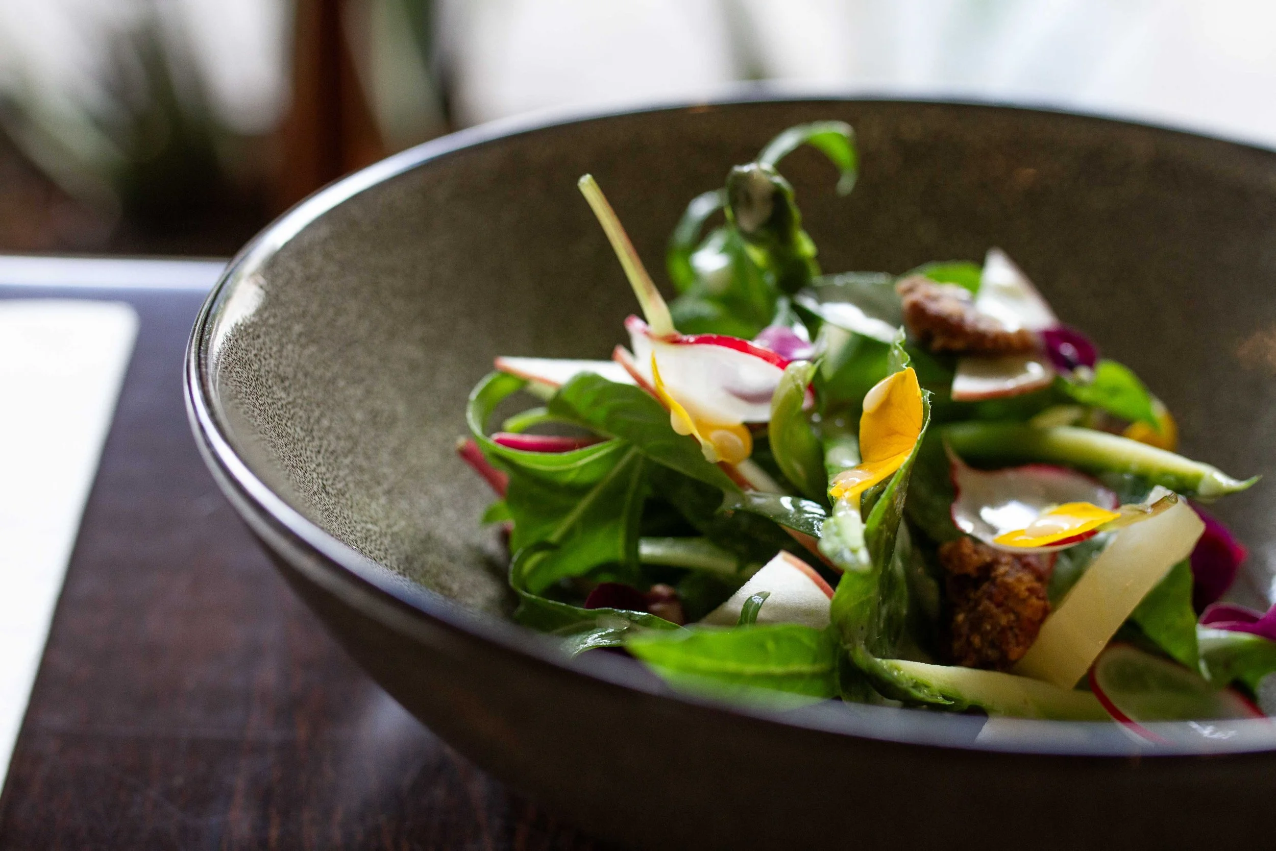 Salad in a dark ceramic bowl with greens, radish slices, cheese, and croutons.