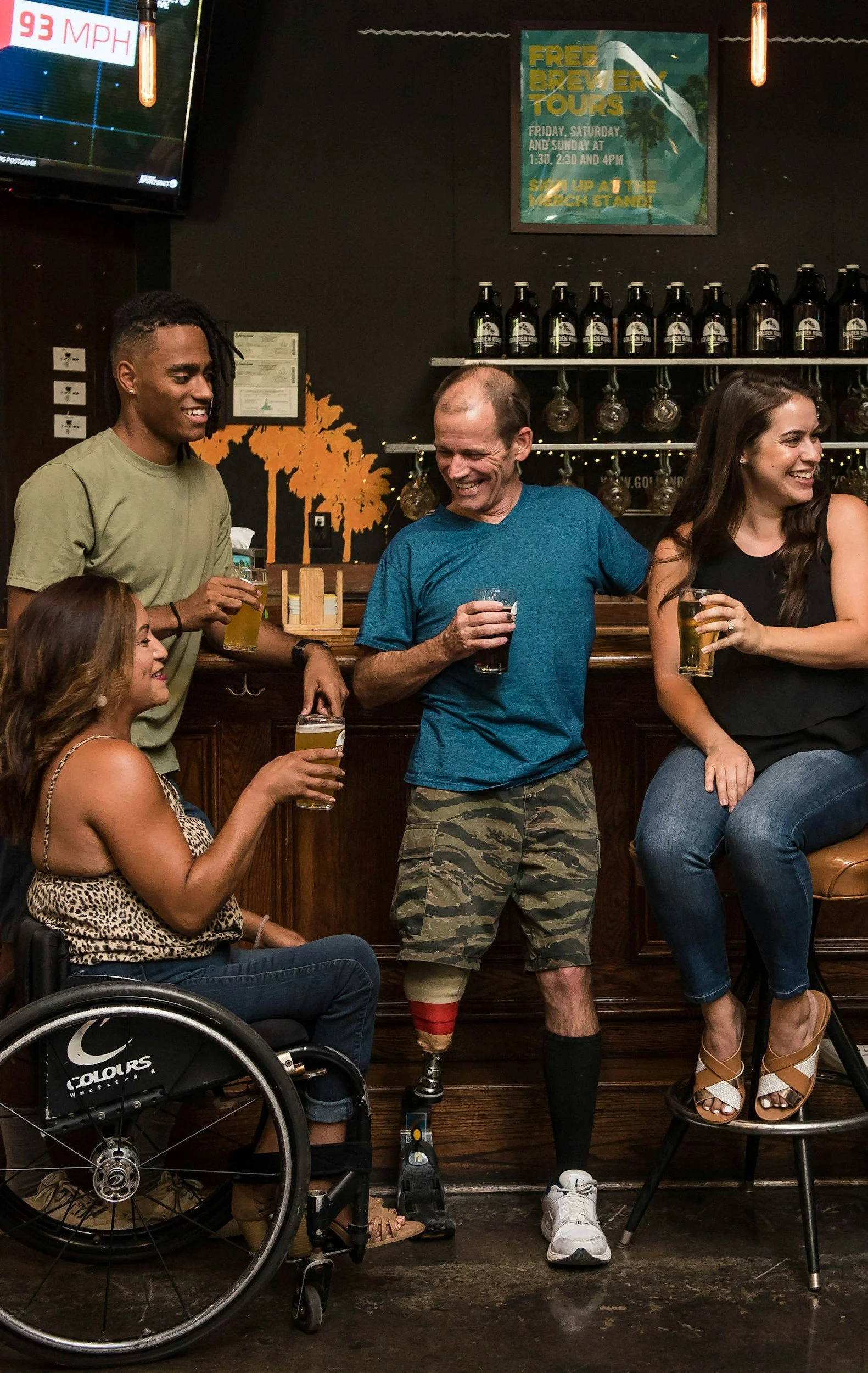 A diverse group of four friends enjoying drinks at a bar, one person in a wheelchair and another with a prosthetic leg. They are smiling and engaged in conversation.