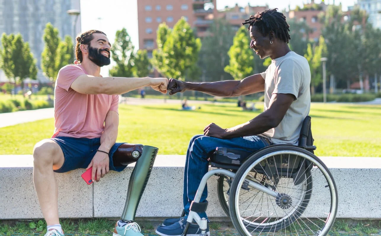 Two men interact outdoors; one with a prosthetic leg sitting on a concrete bench, the other in a wheelchair. They are exchanging a fist bump in a park setting.