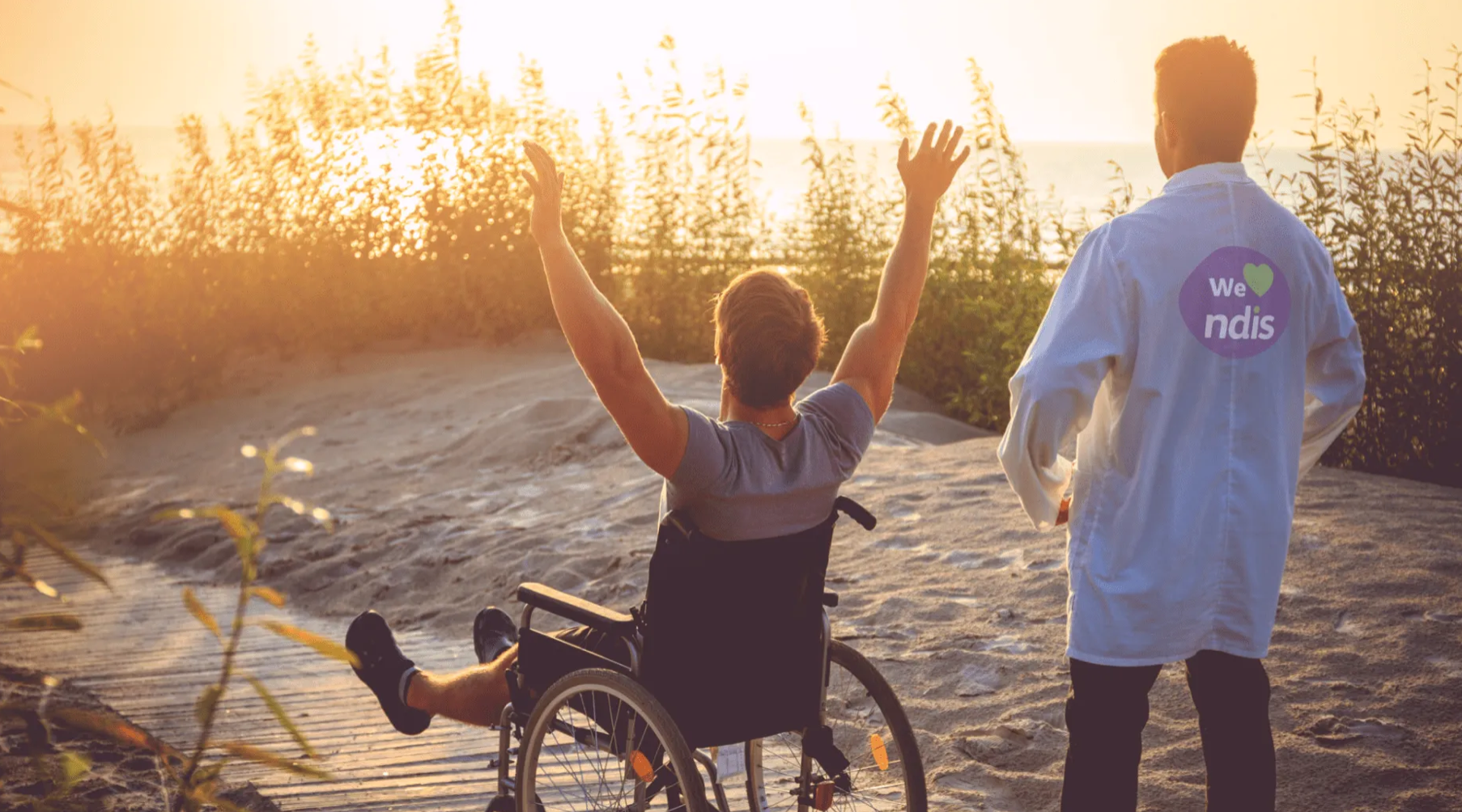 A person in a wheelchair with arms raised facing the sunset on a sandy path, accompanied by a person with a