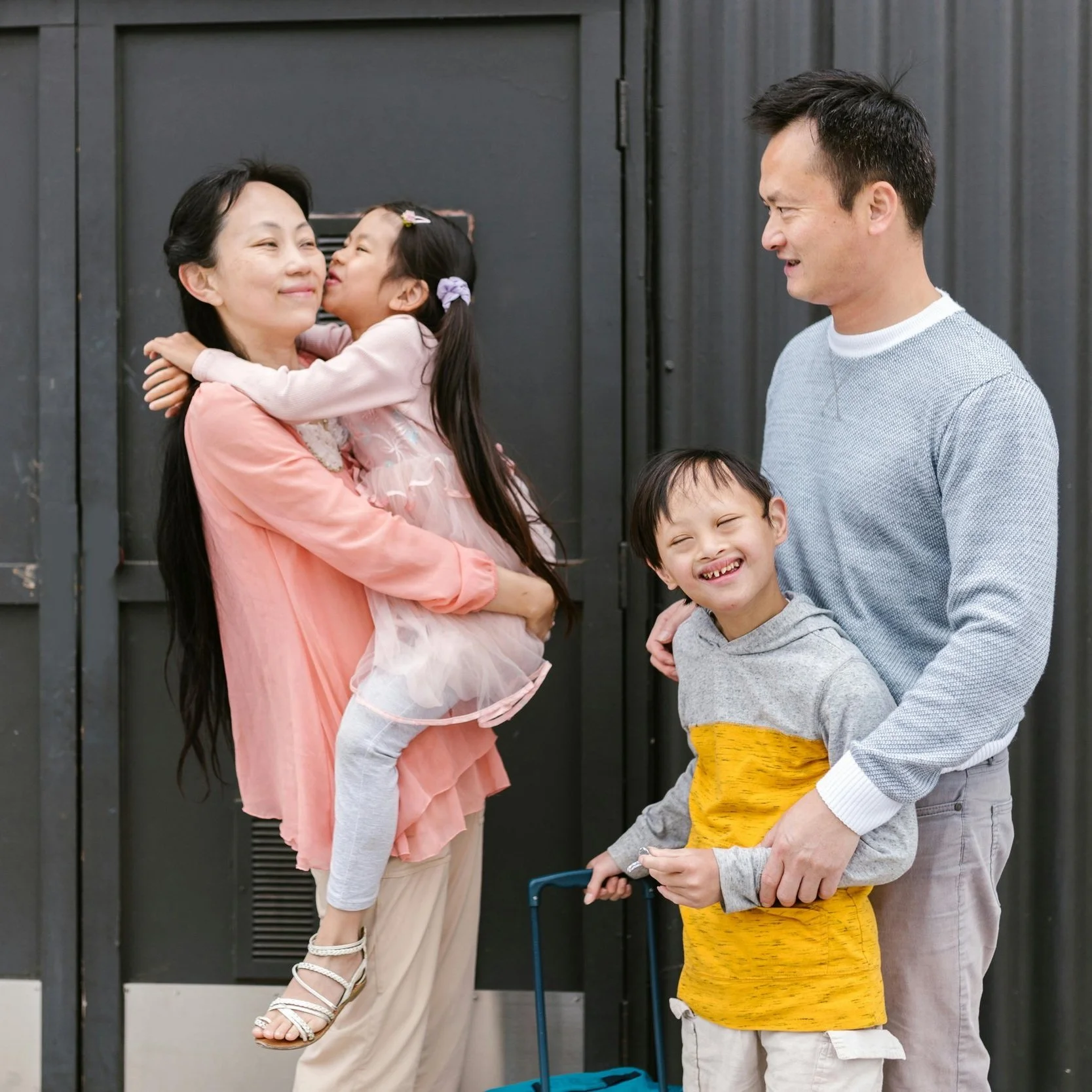 A family of four standing together smiling. The mother is holding a young girl who is hugging her. The father is standing next to a boy holding a suitcase and smiling joyful. They are outdoors in front of a dark door.