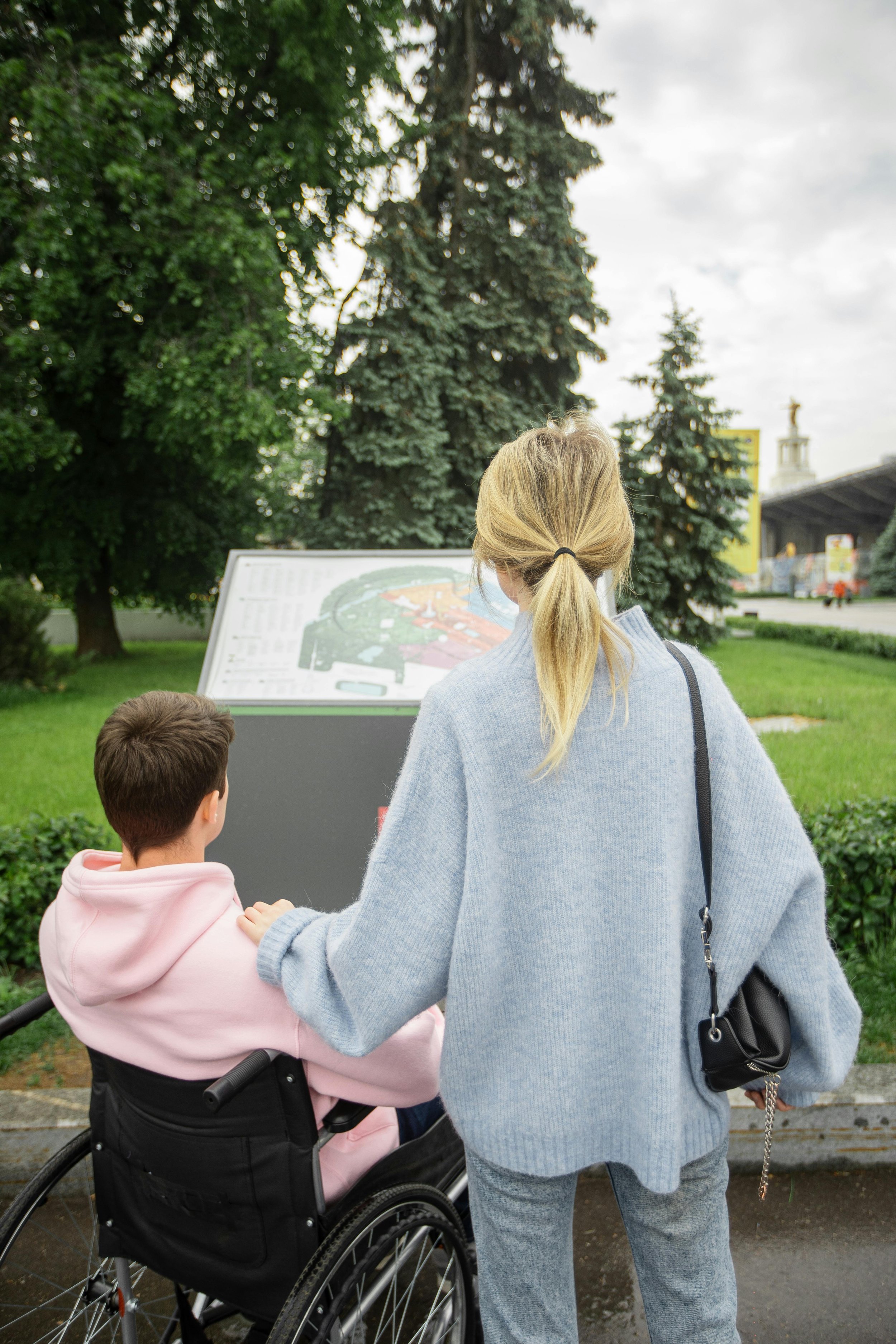 A person in a wheelchair wearing a pink hoodie is accompanied by someone in a blue sweater, looking at an outdoor sign with trees and grass surrounding them.