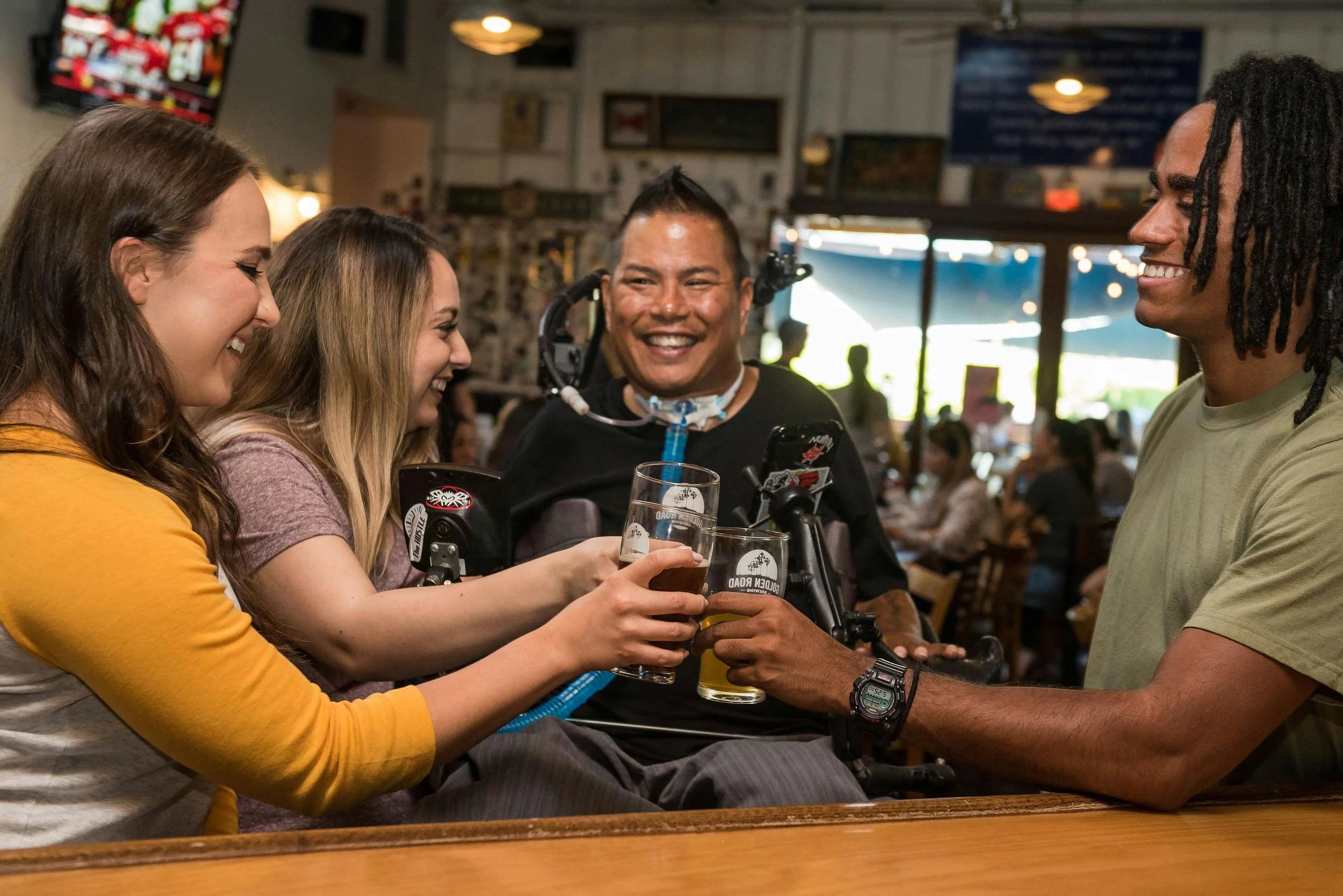 Four people, including a person using a wheelchair, smiling and clinking beer glasses in a bar setting.