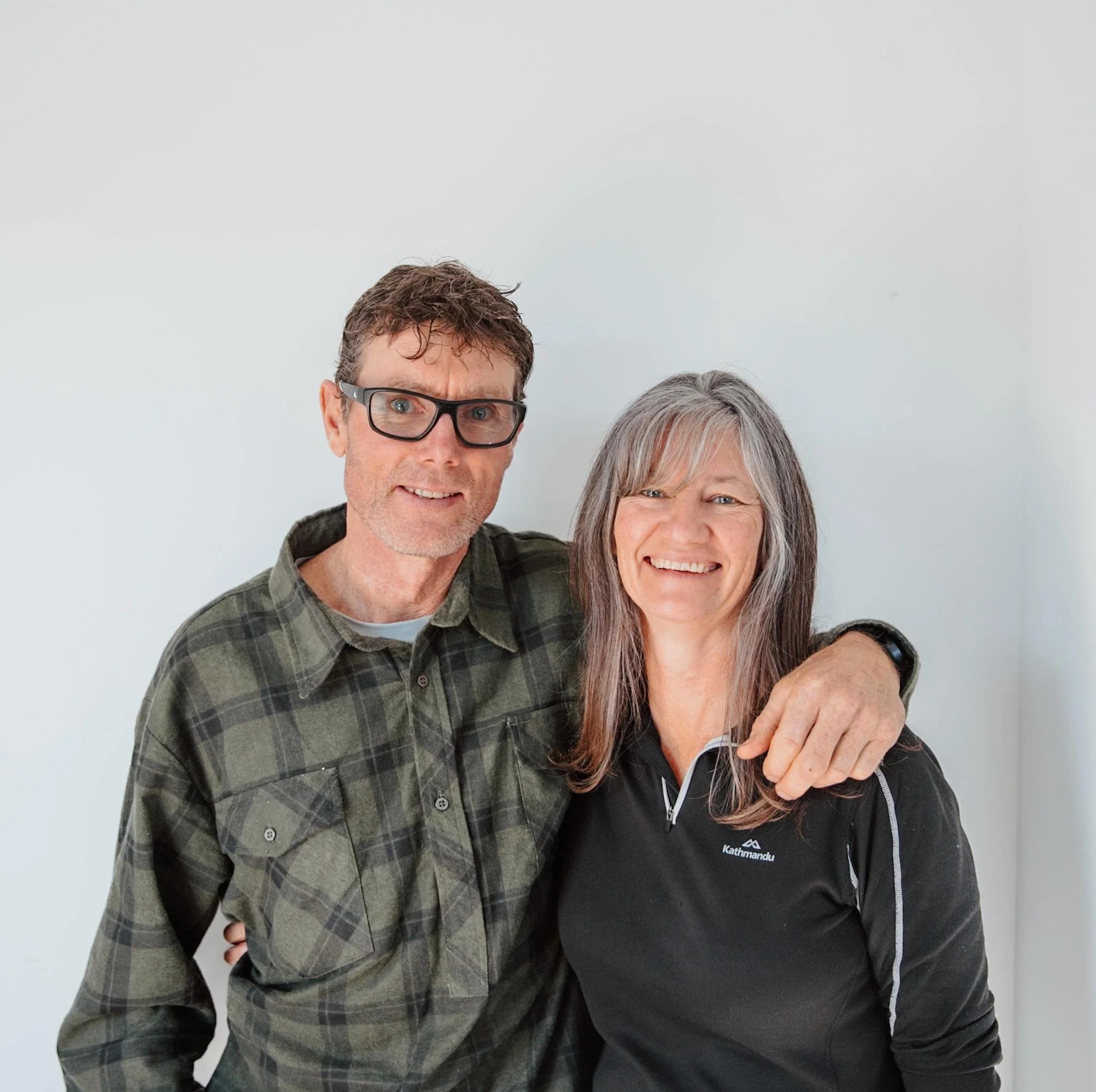 A smiling middle-aged man and woman standing close together, with the man's arm around the woman's shoulder, in front of a plain white wall.