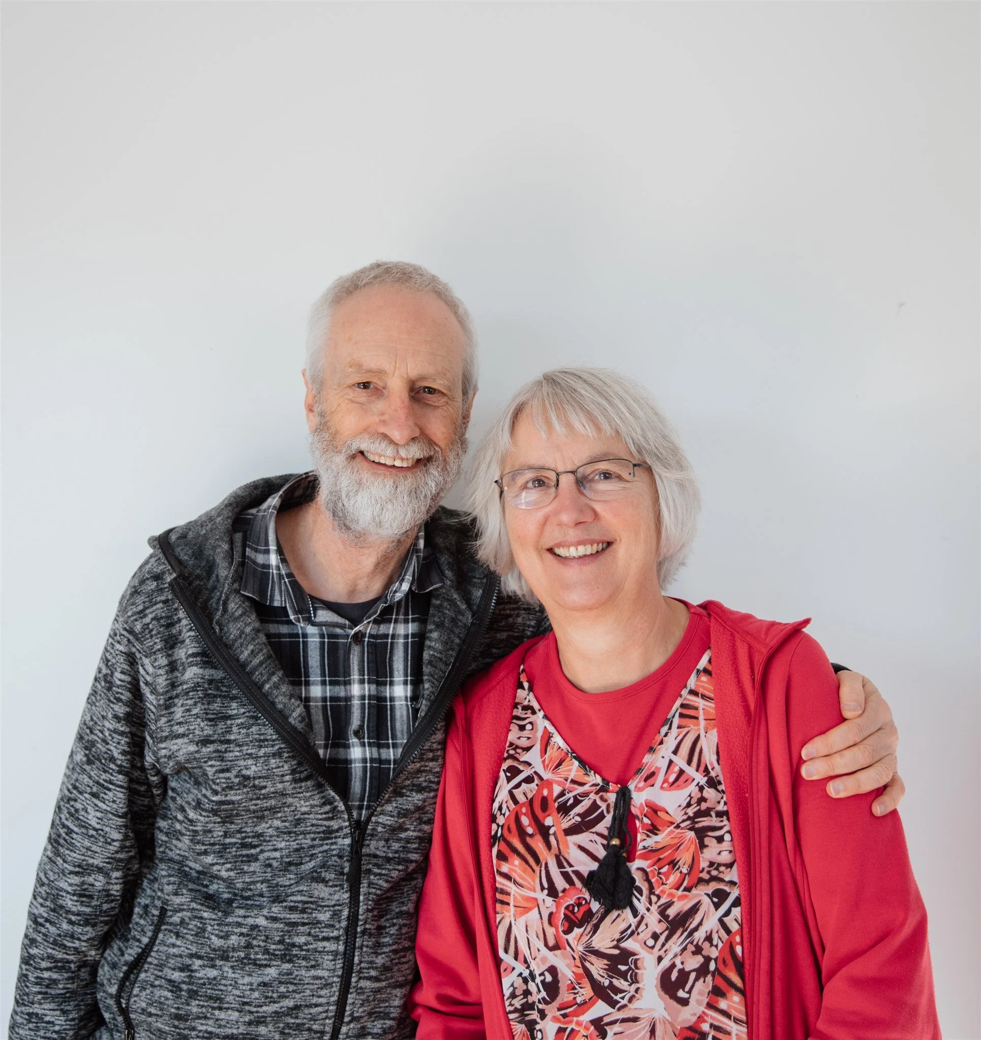 Smiling elderly man and woman standing close together, with the man wearing a gray zip-up hoodie and black plaid shirt, and the woman wearing glasses, a red jacket, and a patterned shirt, against a plain white background.