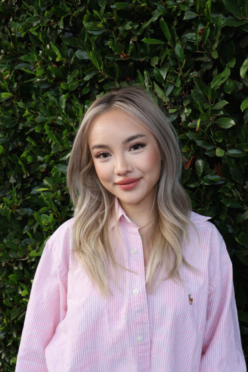 A young woman with blonde hair and a pink striped shirt standing in front of green leafy bushes, smiling at the camera.