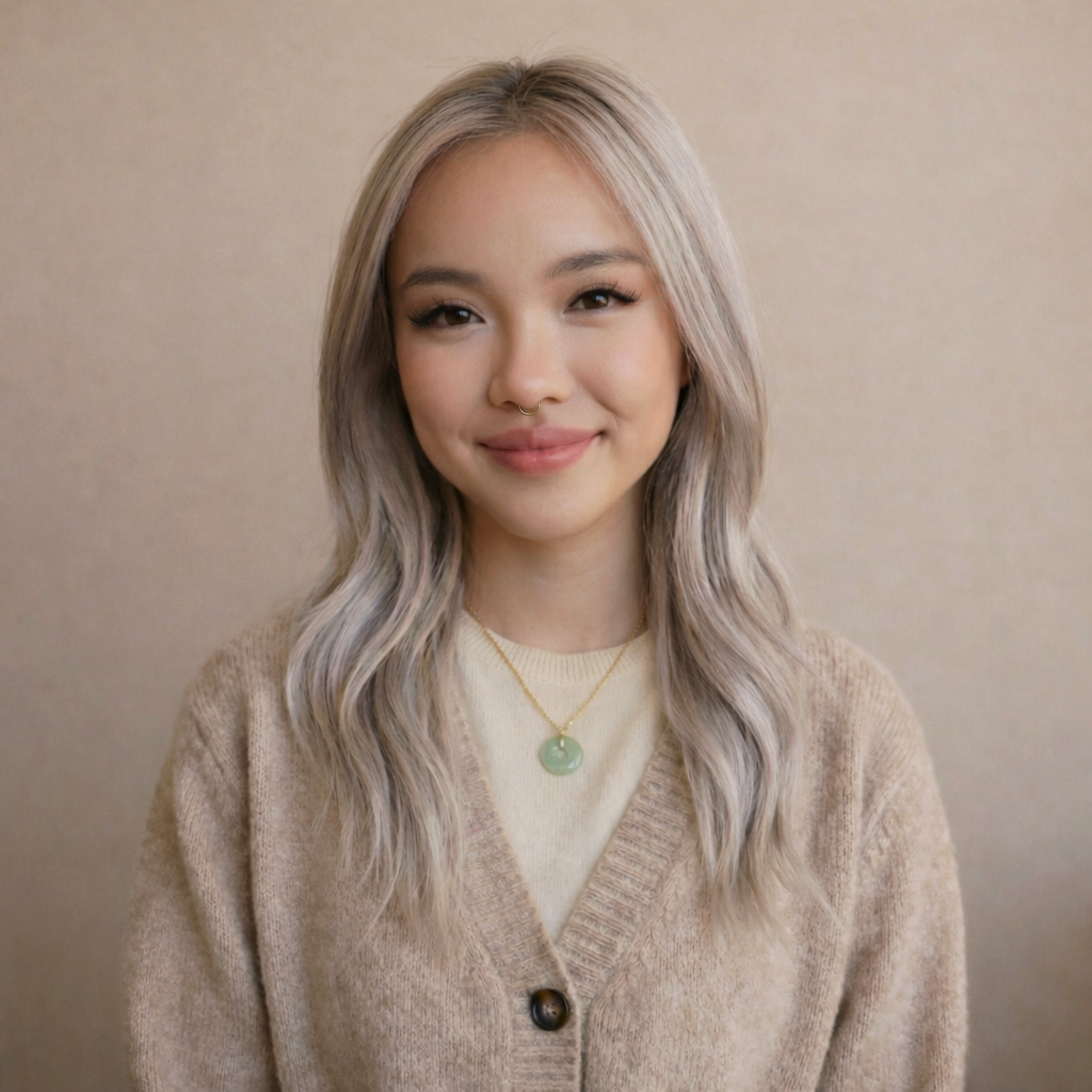 Portrait of a young woman with blonde wavy hair, light skin, wearing a beige cardigan over a cream top, and a green jade pendant necklace, standing in front of a neutral beige background.