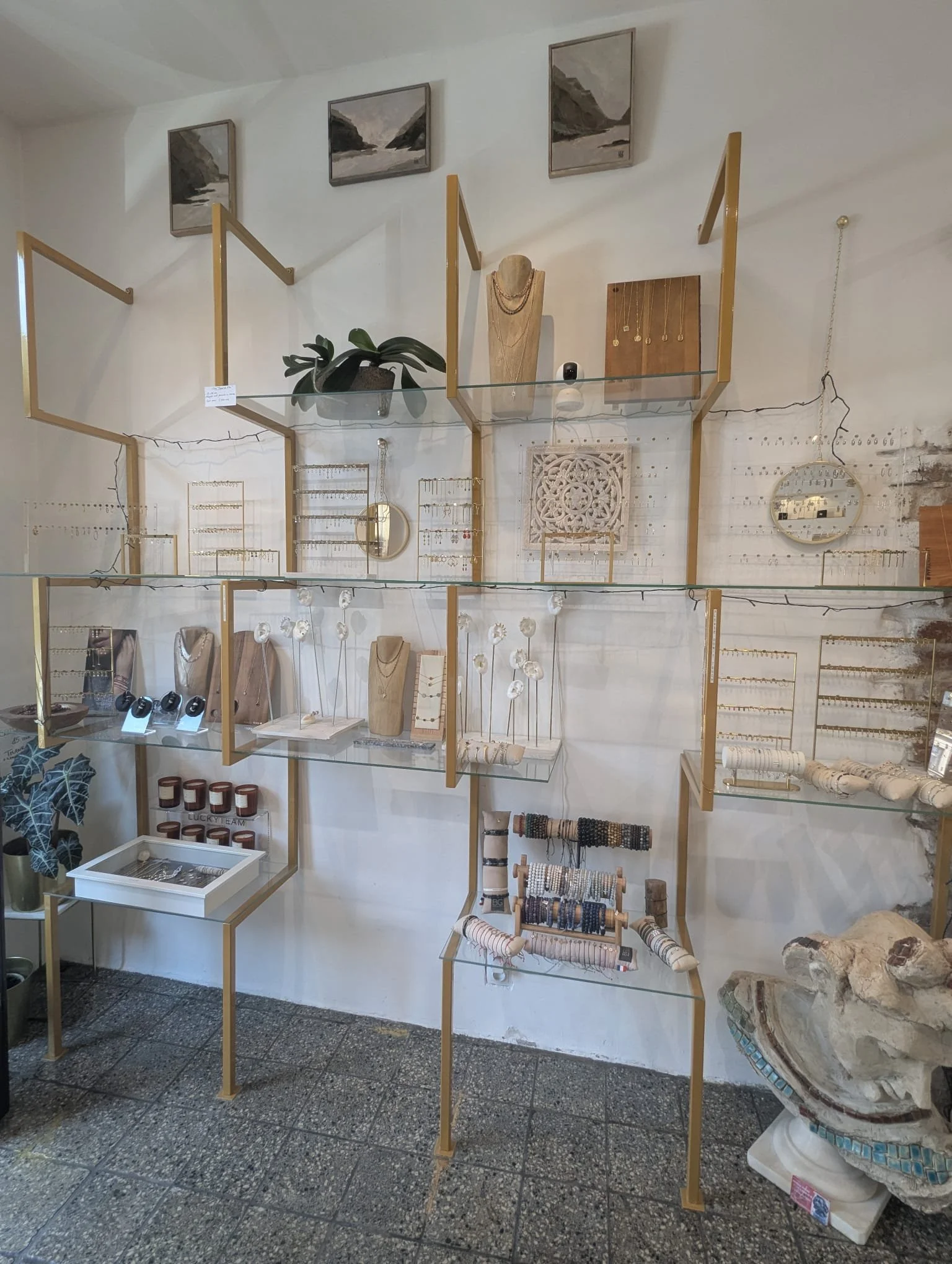 Display of jewelry and decorative items on glass and wooden stands against a white wall with three landscape paintings and a tiger sculpture.