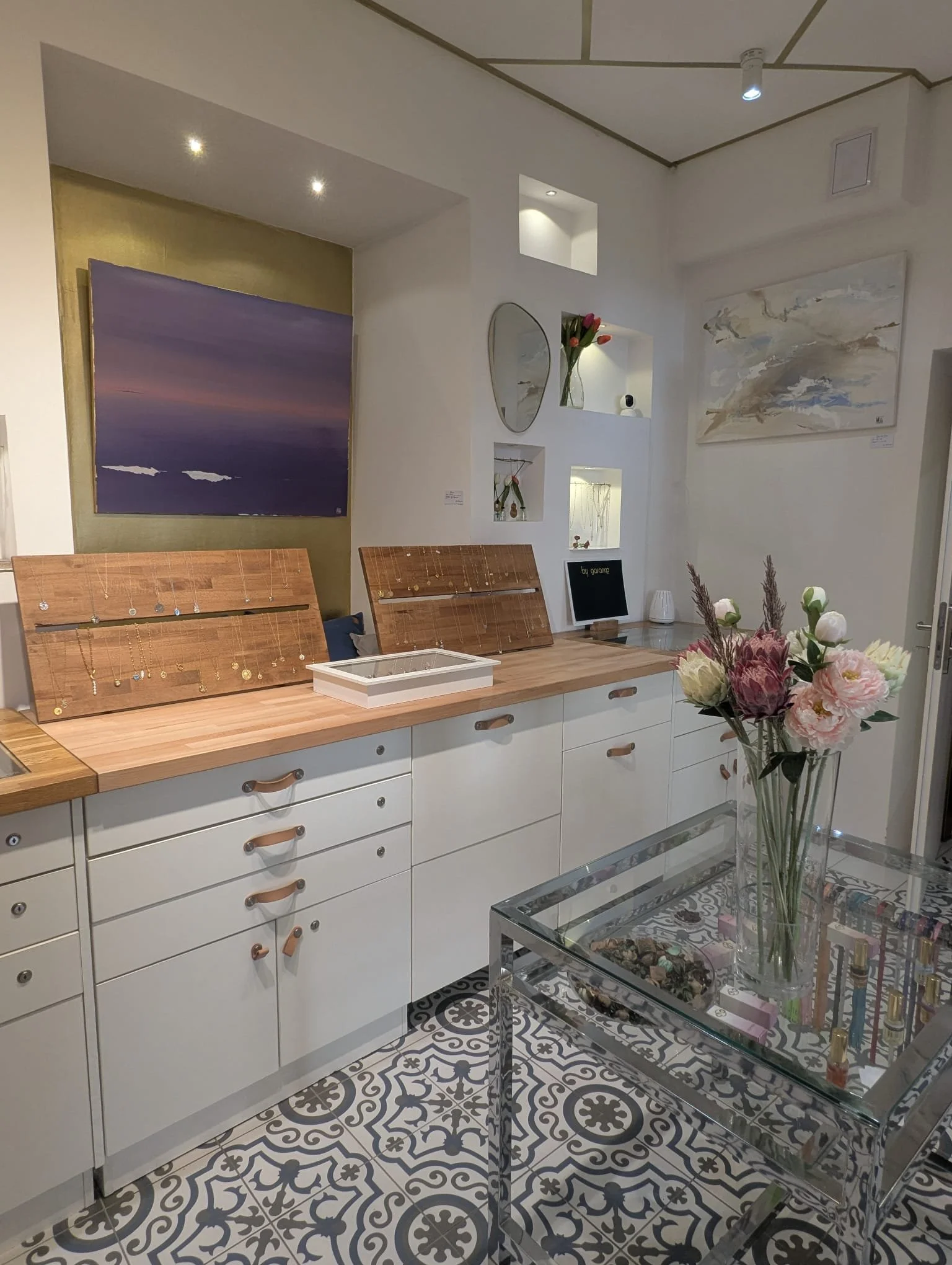 Interior art gallery and jewel shop whith white cabinet, glass-topped table with flowers, wall art and jewelry display.