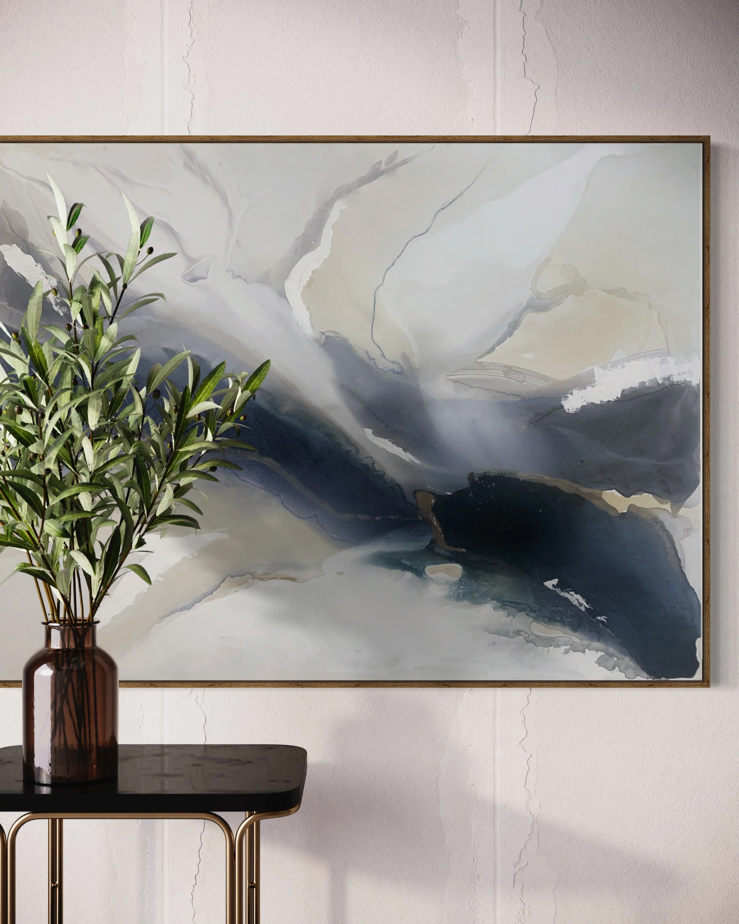 A tall, brown glass vase with green leafy plants sits on a black table with gold legs, positioned next to a framed abstract artwork with shades of gray, black, and white, on a white wall with faint cracks.
