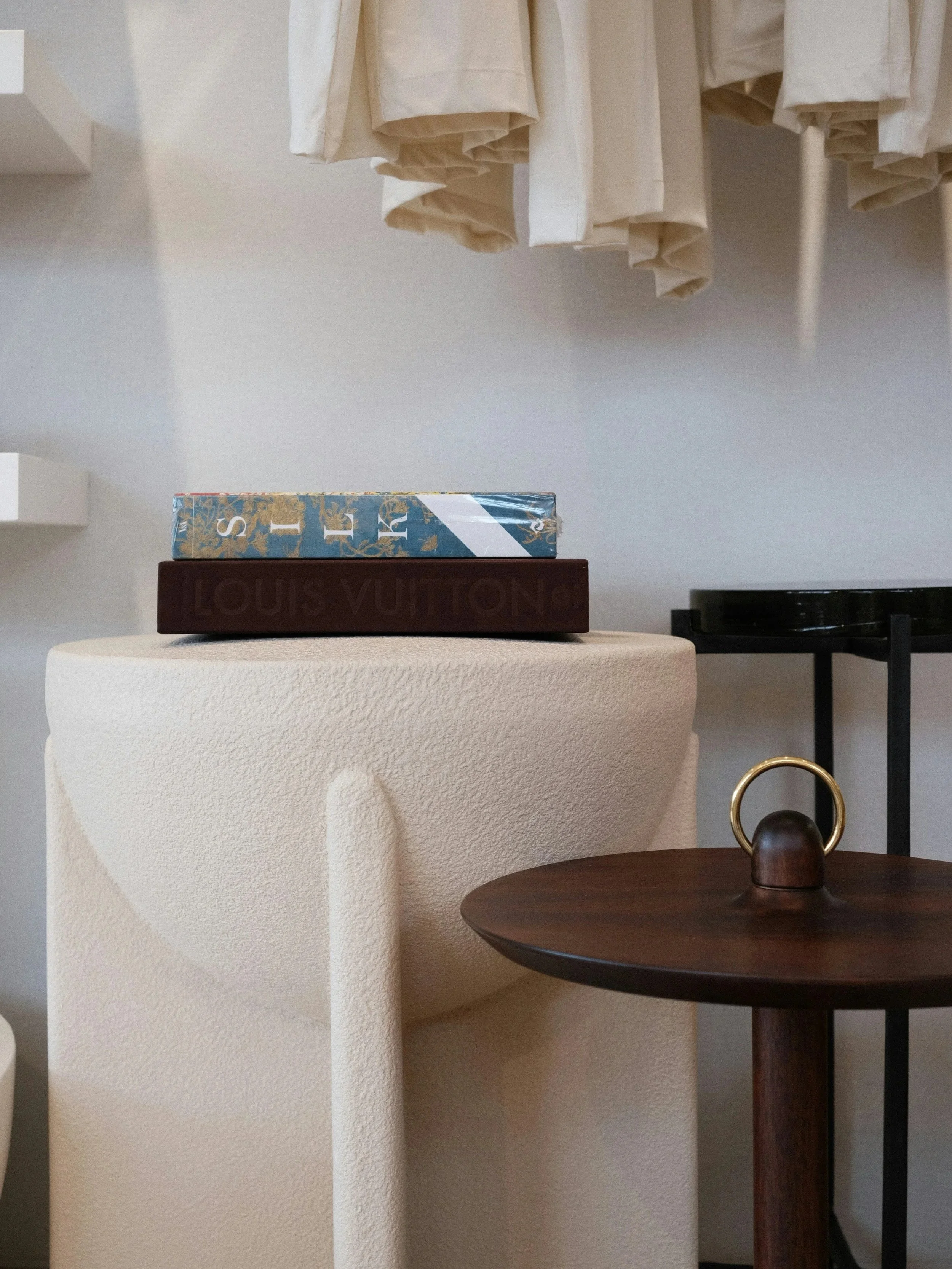 Minimalist wardrobe layout, three stacked boxes on a cream-colored textured pedestal, beige curtains hanging above, a dark wooden side table with a decorative brass ring, and a black object on the table, with white walls in the background.