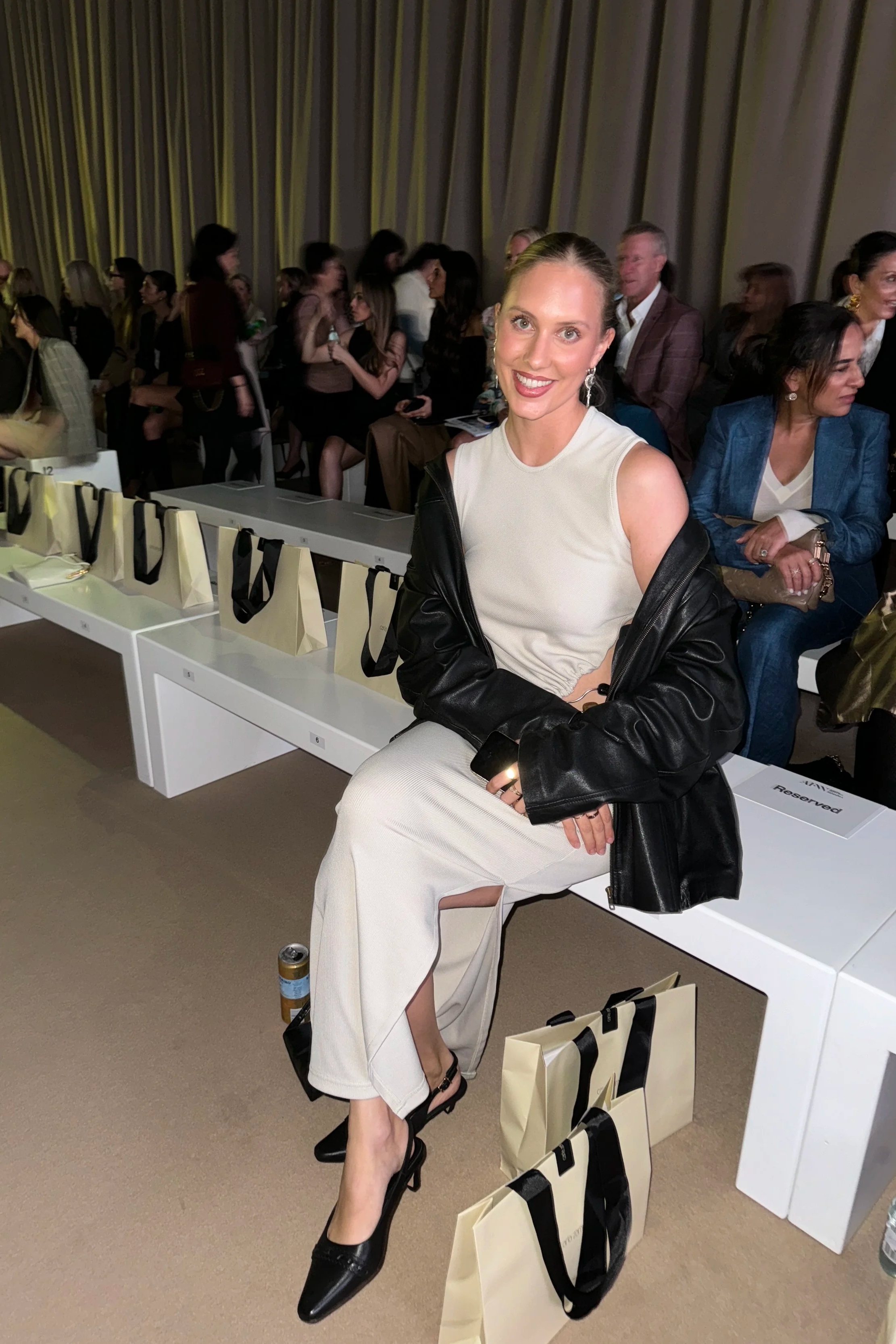 A woman in a white dress and black high heels sitting on a chair with her legs crossed, smiling, with several shopping bags on the floor and sitting on a white bench in a room with grey curtains and many people seated behind her.