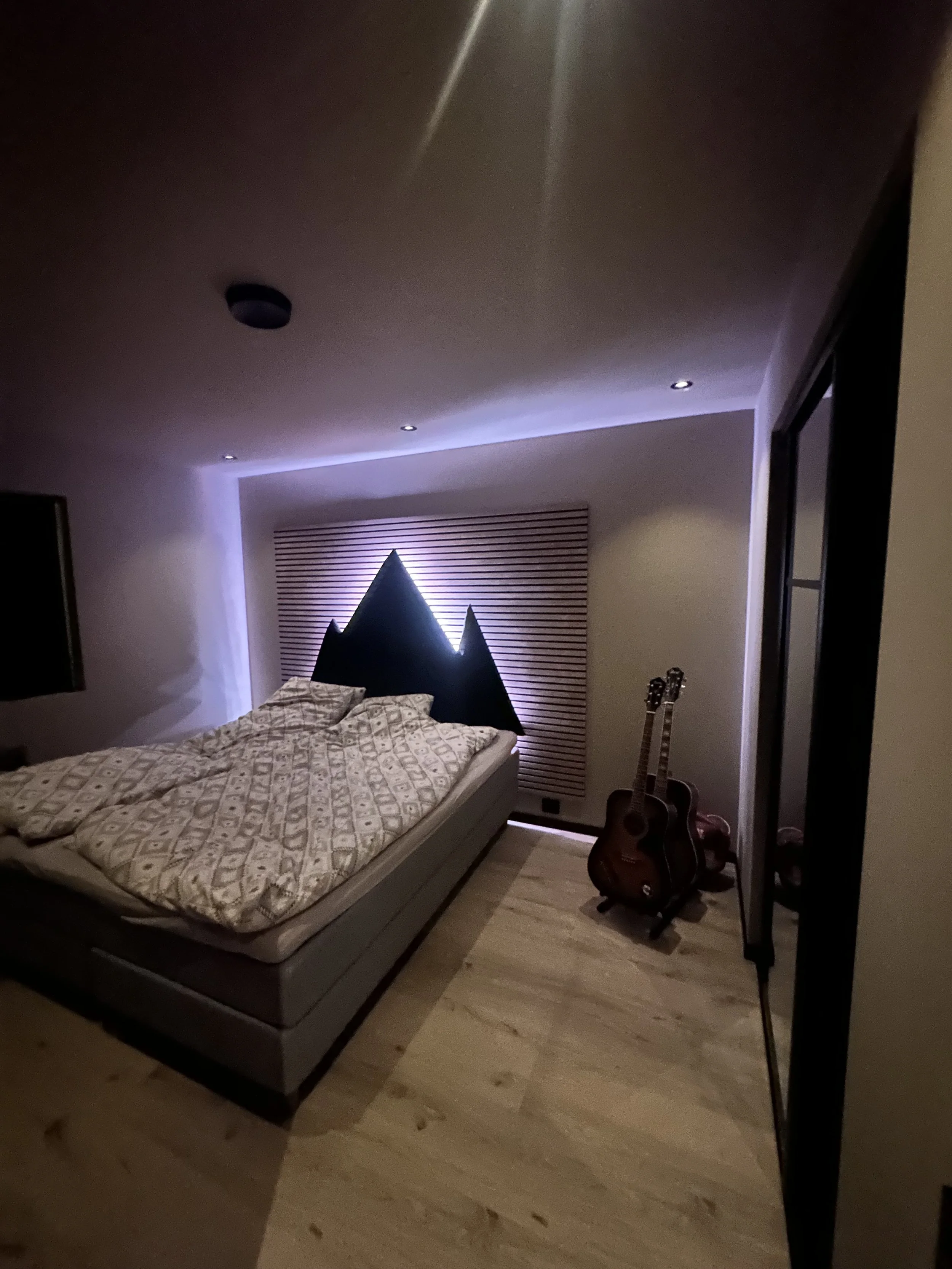 Et soverom med stor seng, to akustikkgitarer ved siden av sengen, og et veggdekor som ser ut som fjell, opplyst bak.