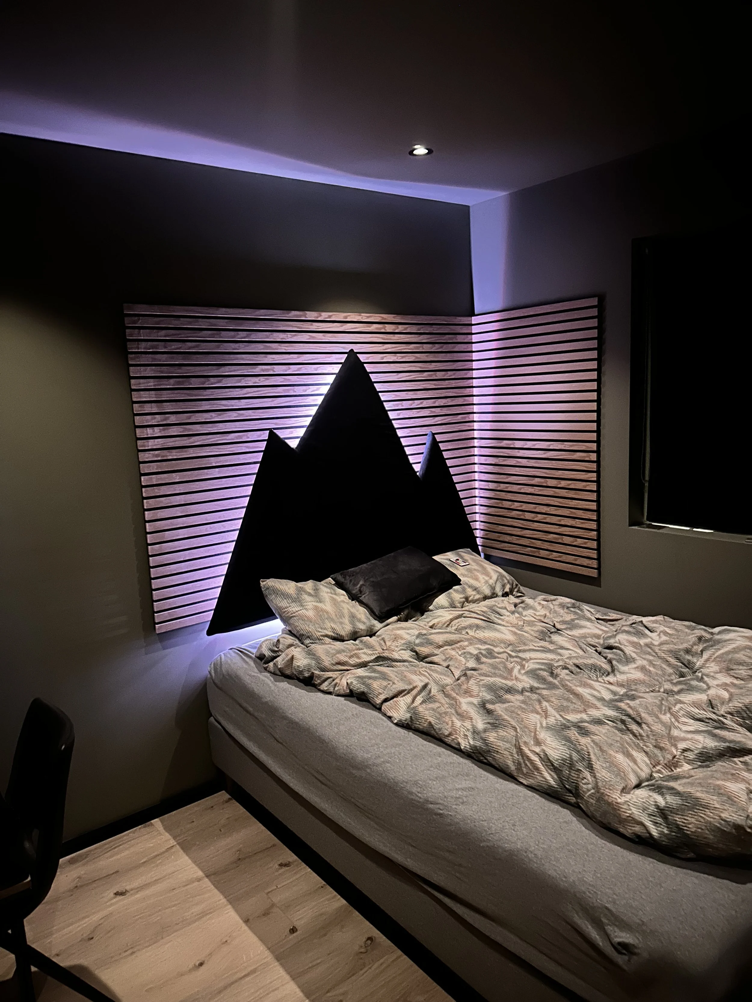 Et soverom med en seng, sengelaken og pute, dekorert med en kunstnerisk hodegavl av fjellformede trepaneler med belysning bak, veggen er malt grå, og det er delvis synlig et vindu på høyre side.