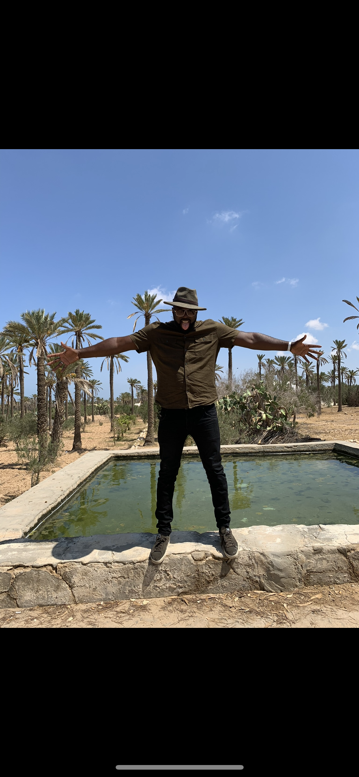 Un homme portant un chapeau, des lunettes et une chemise marron se tient avec les bras ouverts devant une fontaine en pierre, avec un paysage désertique et des palmiers sous un ciel bleu clair.