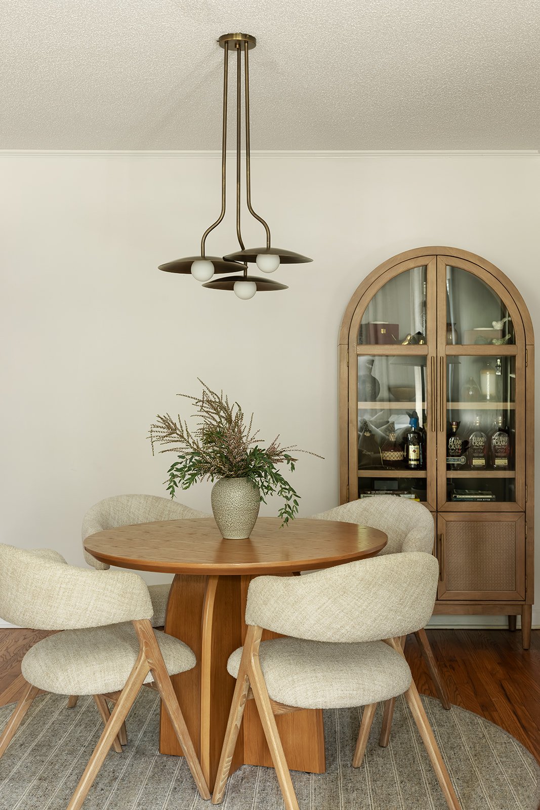A dining room with a round wooden table and four upholstered chairs with light-colored fabric. On the table, there's a vase with greenery and flowers. In the background, a wooden display cabinet with glass doors contains bottles and decorative items. A modern chandelier hangs from the ceiling, and the room has neutral-colored walls and wooden flooring.