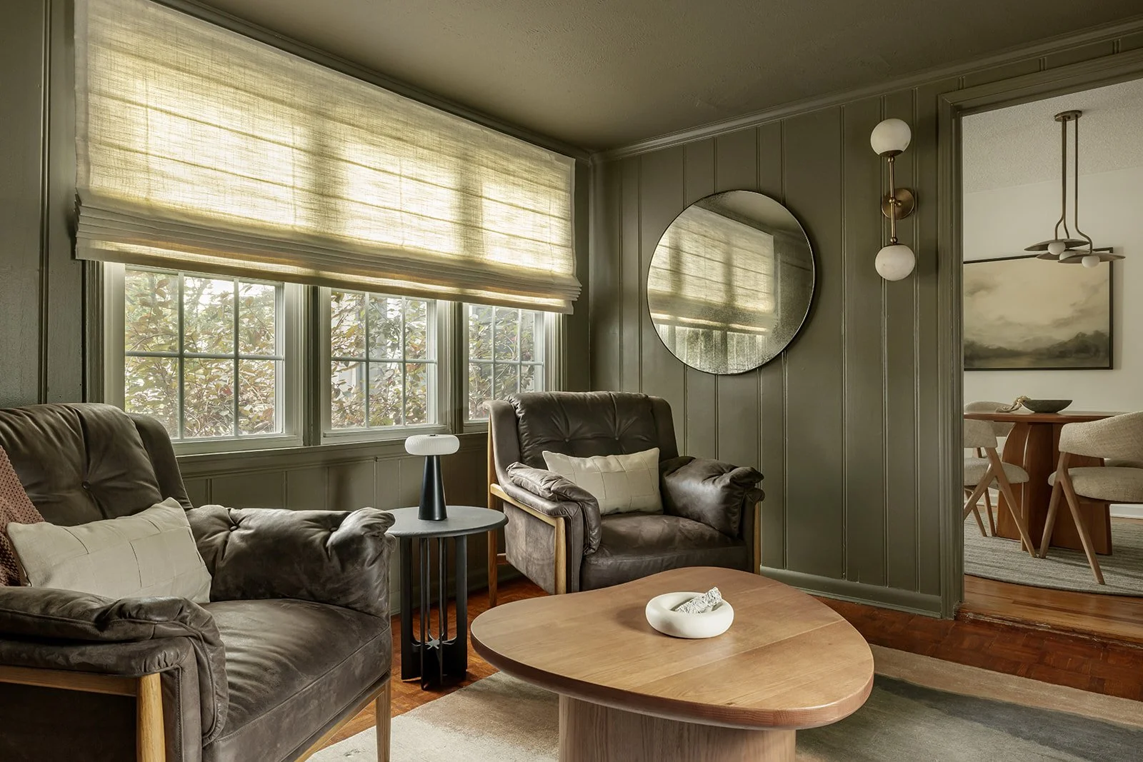 Living room with large windows covered by beige blinds, featuring two brown leather armchairs with cream pillows, a wooden coffee table, a side table with a modern black lamp, an oval mirror on the green paneled wall, and a view into a dining area with a round table and chairs.