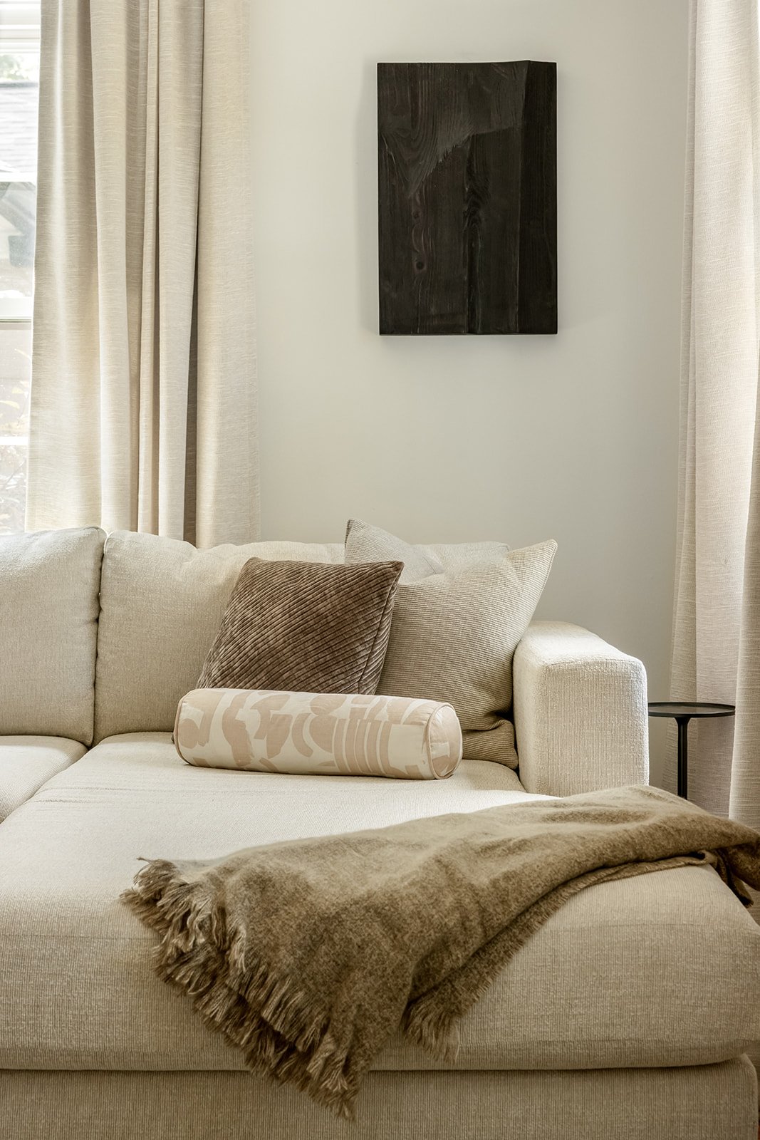 Living room with a beige sectional sofa decorated with pillows and a throw blanket, beige curtains, a dark wall art piece, and a small side table.