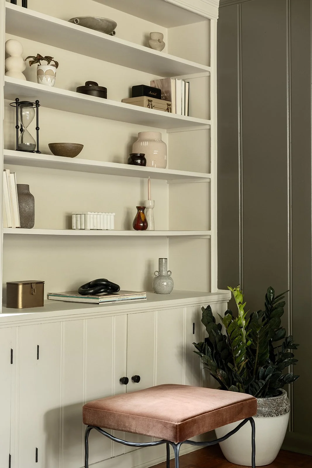 Interior view of a built-in white bookshelf with decorative items, books, and a potted plant nearby, and a pink velvet stool in front.