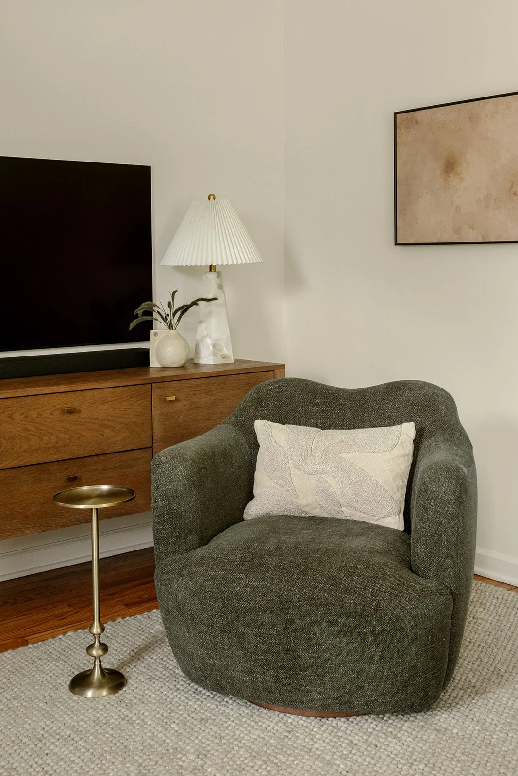 A living room corner with a dark grey armchair with a decorative pillow, a wooden dresser with a bookshelf, a flat-screen TV, a white table lamp, a white vase with plant, and a framed beige abstract artwork on the wall.