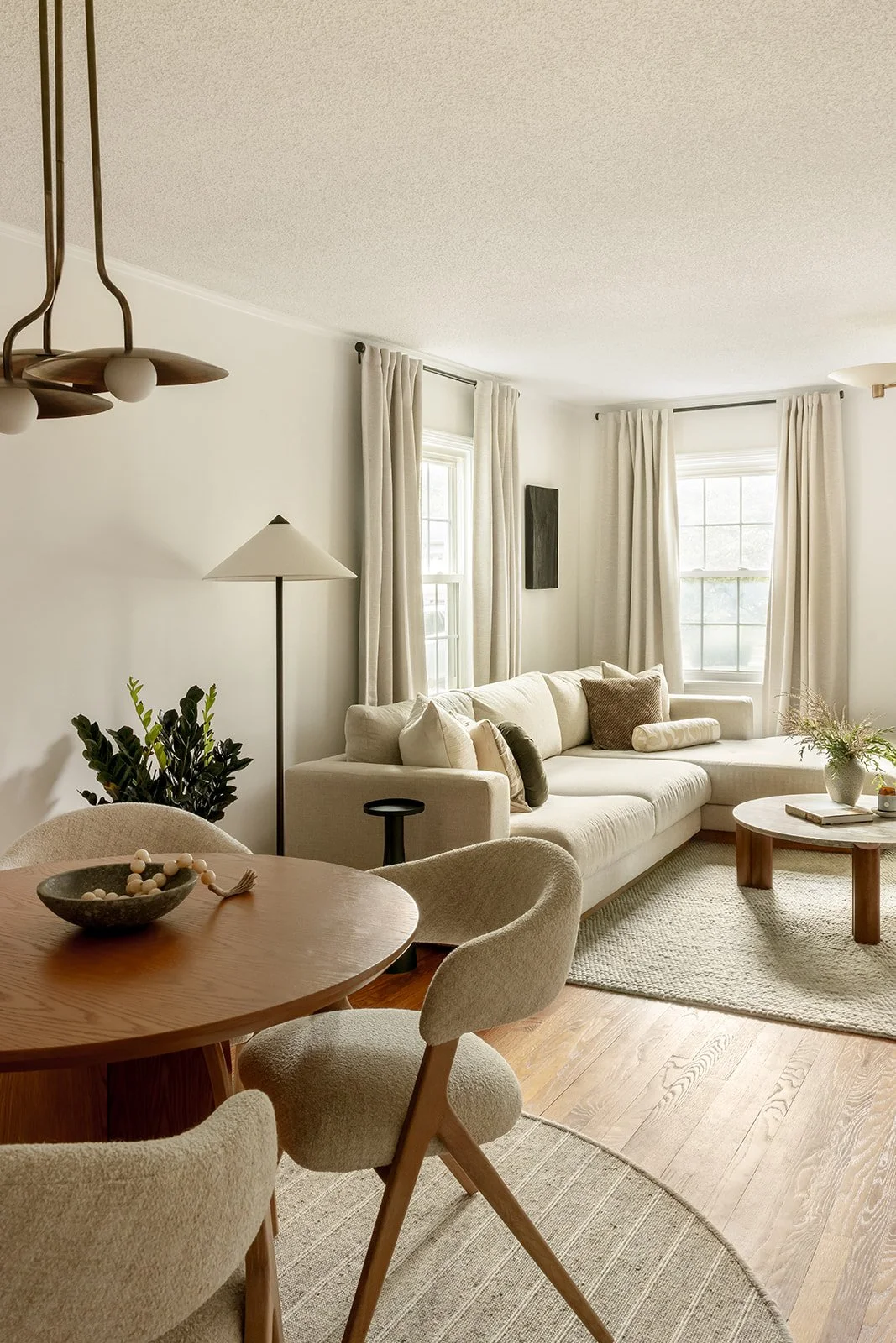 Living room with a beige sectional sofa, white walls, and wooden floors. There are two large windows with white curtains, a white floor lamp, a potted plant, and a round wooden coffee table. A dining table with beige chairs is visible in the foreground.