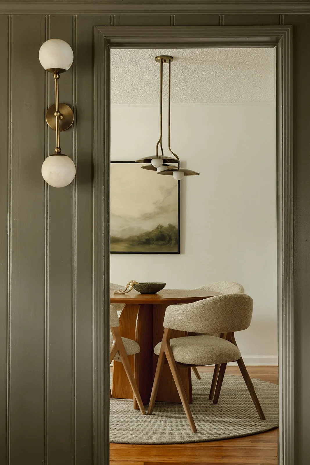 View through doorway into dining room with wooden table, beige upholstered chairs, a modern chandelier, wall art, and a decorative bowl on the table.