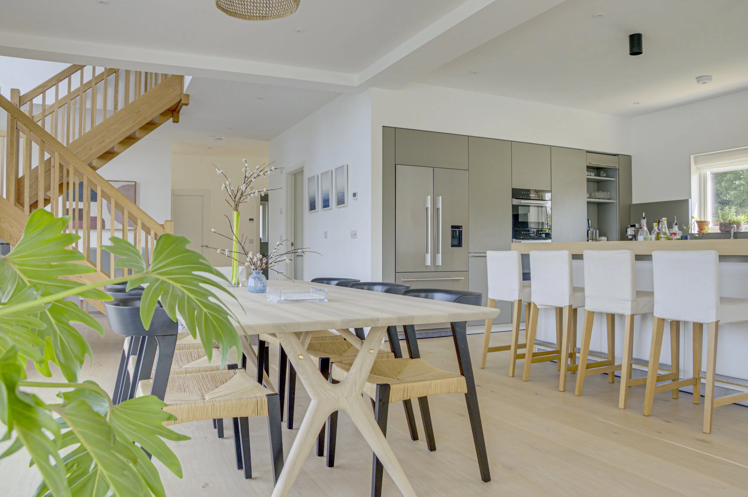 Modern open-concept kitchen and dining area with light wood flooring, a large wooden dining table with black and natural wood chairs, a kitchen bar with white bar stools, stainless steel appliances, a small window with potted plants, and a staircase