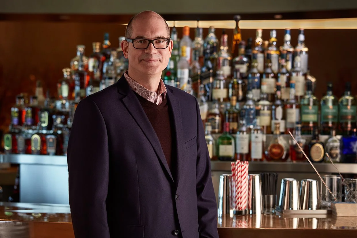 A man in glasses and a dark suit standing behind a bar with bottles of liquor in the background.