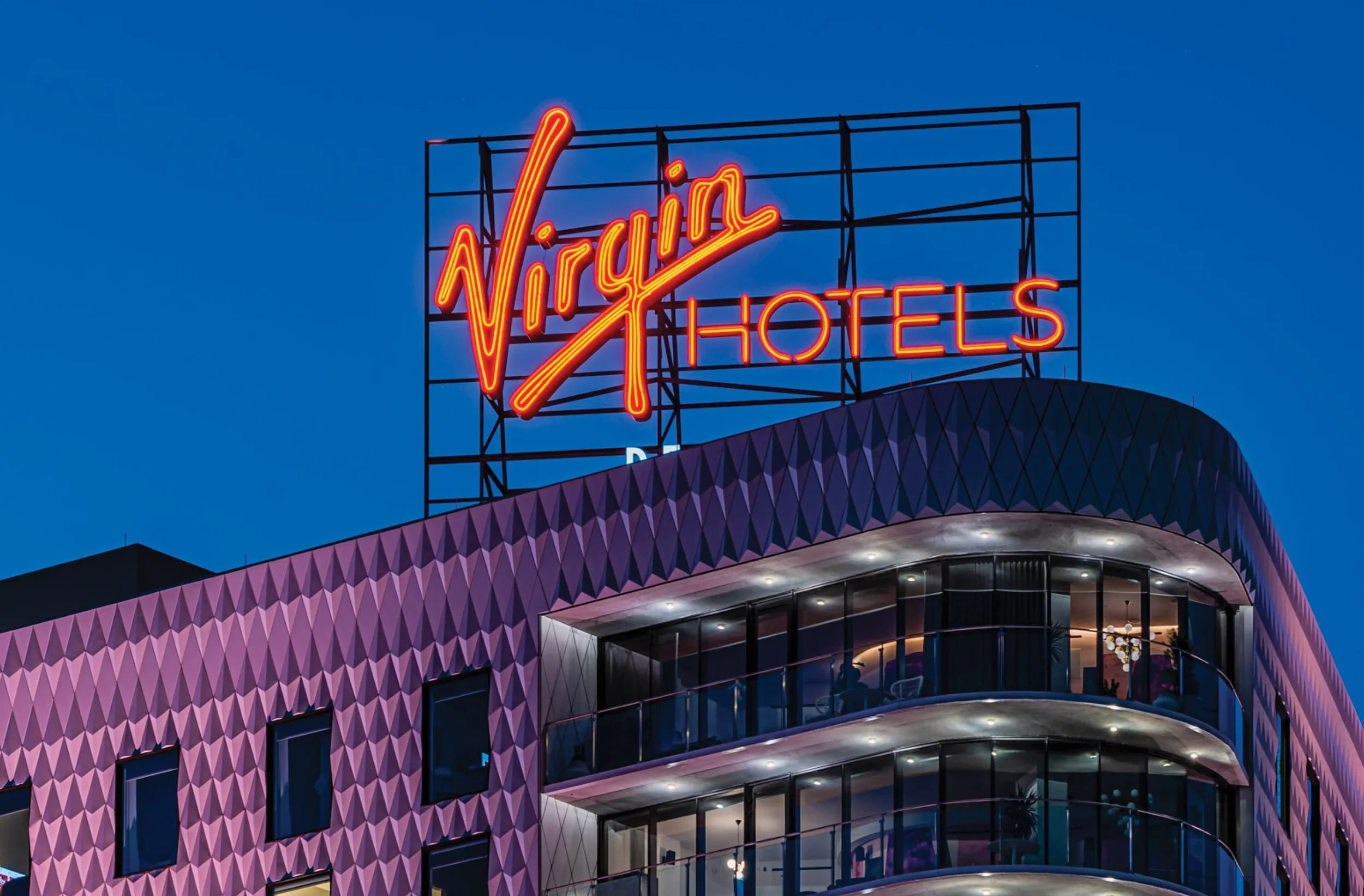 Neon sign reading 'Virgin Hotels' on the top of a modern building with purple geometric exterior and glass balconies, against a dark blue evening sky.