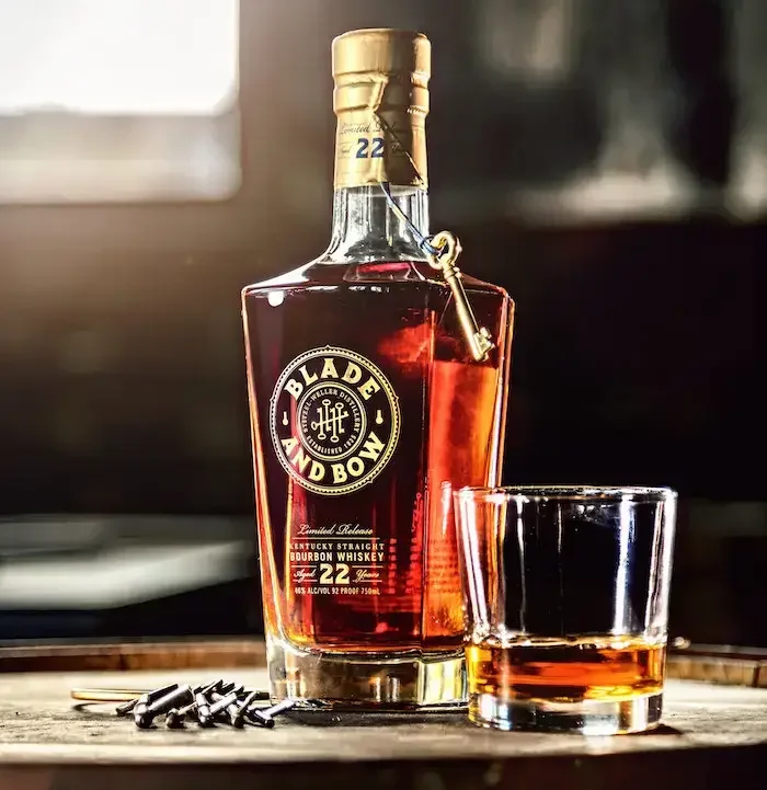 A bottle of Bladland Bond bourbon whiskey with a glass of whiskey next to it on a wooden surface, with coins and a blurred background.