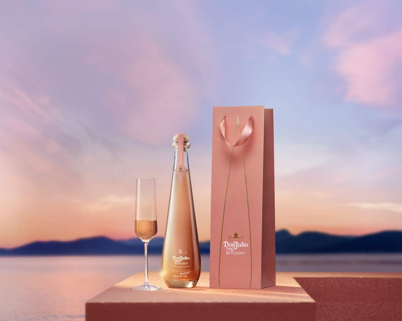 A bottle of Don Julio Rosado tequila and a champagne flute filled with a pink beverage on a pink surface, with a pink gift bag featuring a flower design, set against a sunset sky over a body of water.