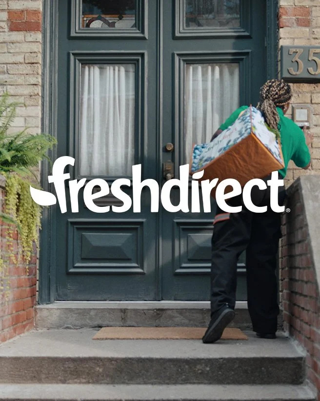 A person wearing a green jacket and black pants carrying a laundry basket into a house through a front door with the FreshDirect logo superimposed.